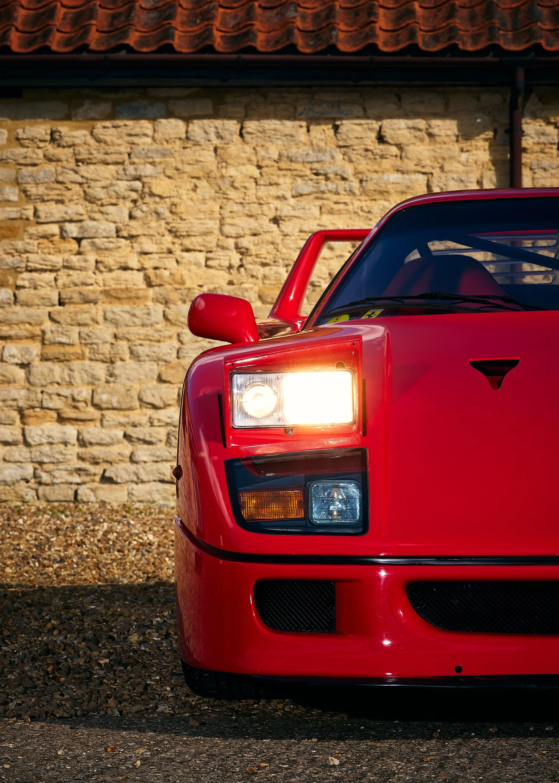 Close-up of the front left corner of a red Ferrari F40 parked on gravel, with a stone wall in the background.