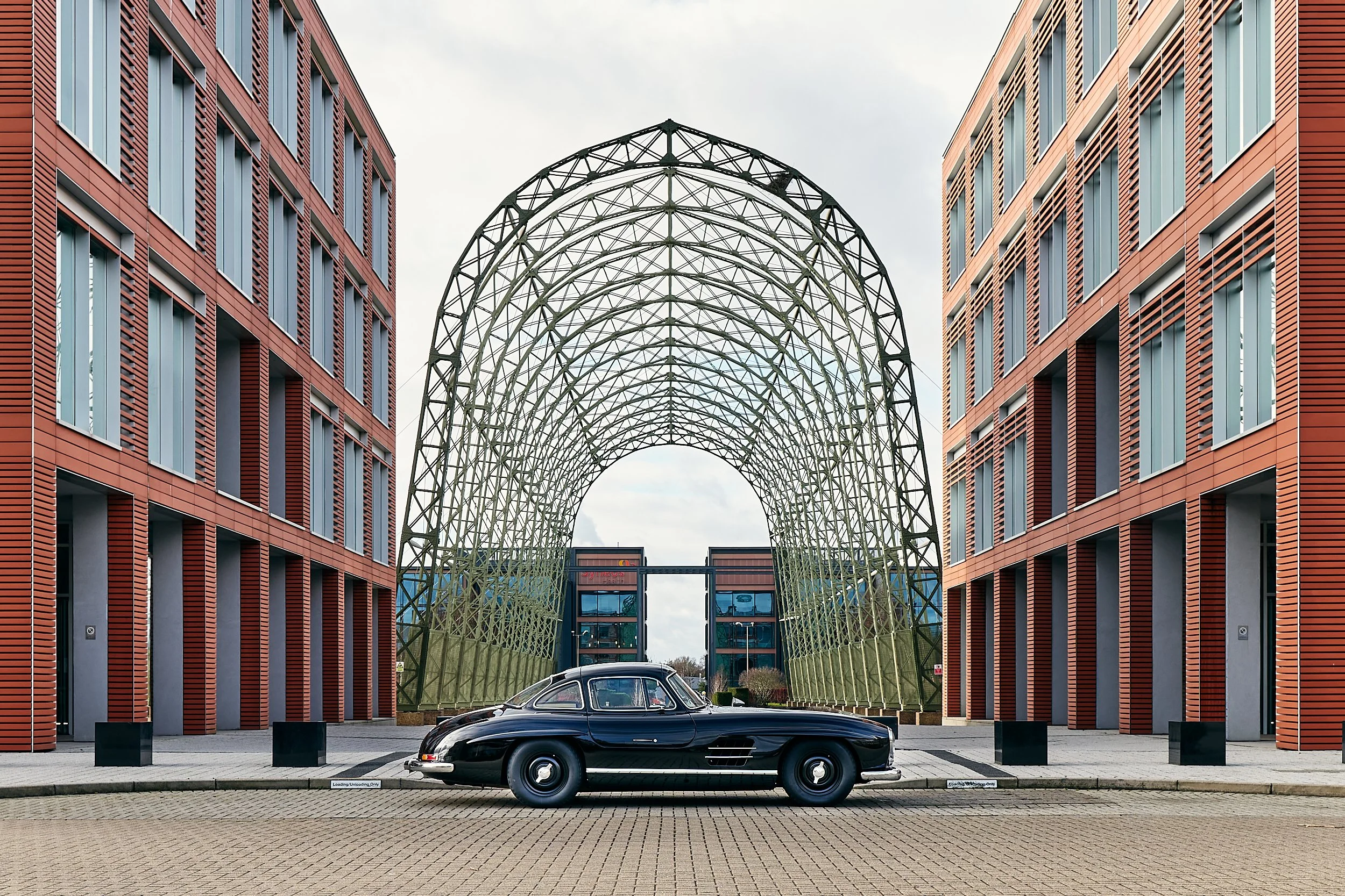 A classic black Mercedes-Benz 300SL Gullwing sports car parked in front of modern red brick buildings with large rectangular windows and metal architectural arches connecting them.