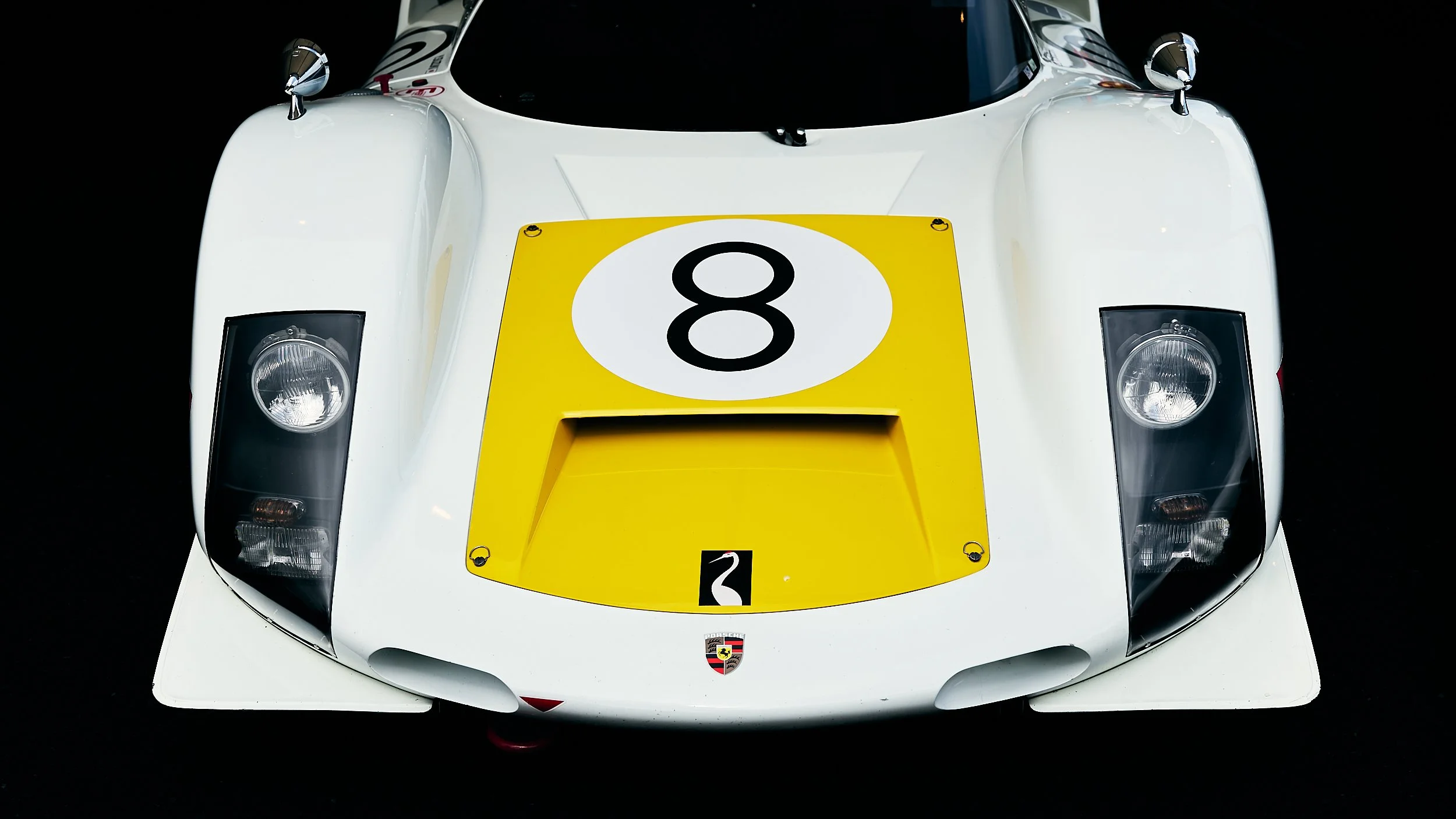 Front view of a white Porsche race car with yellow and black number 8 on the hood, Porsche crest logo at the front bottom, and black headlights.