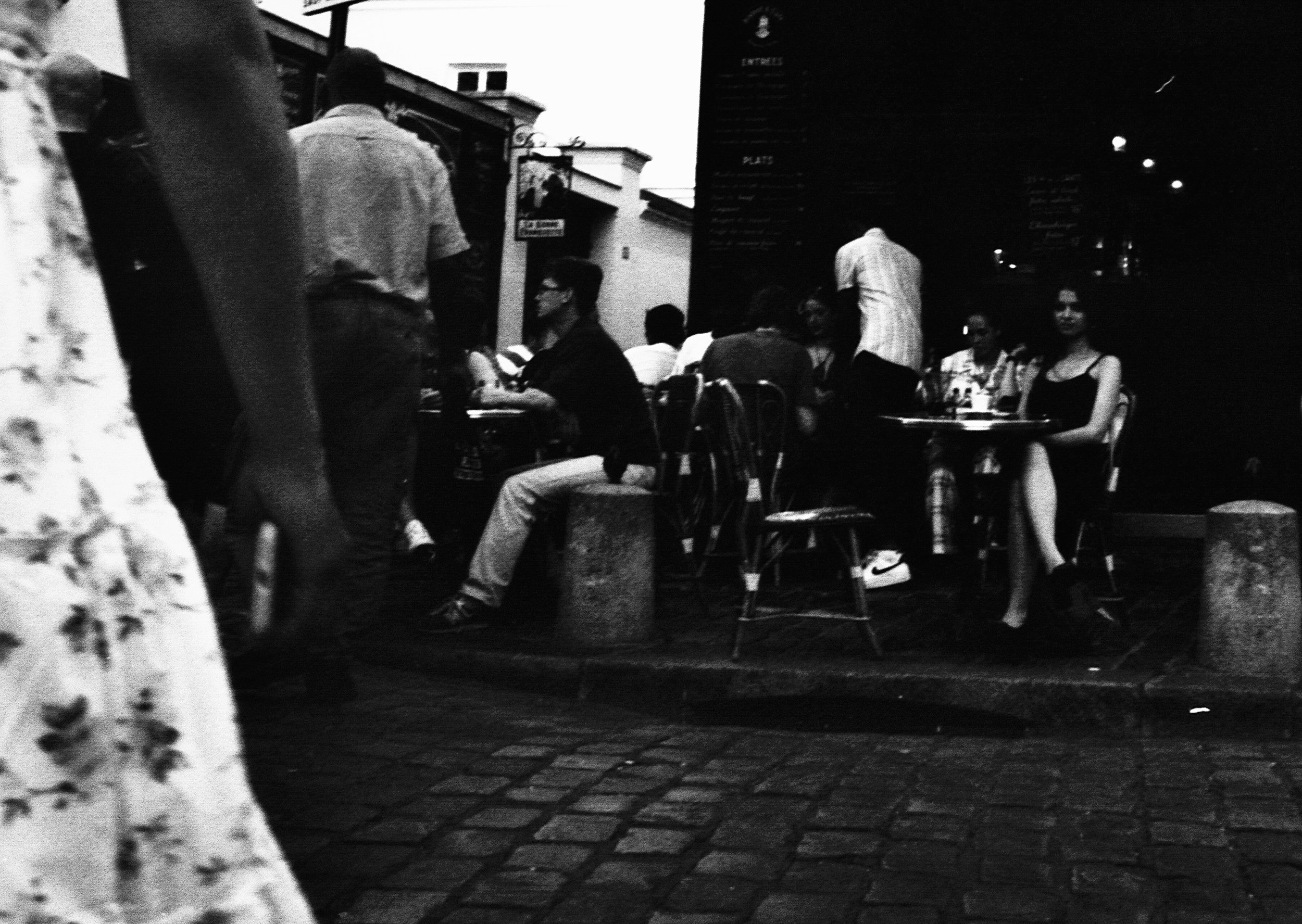 Groupe de personnes assises à une terrasse de café ou restaurant en soirée, avec quelques individus debout, dans un contexte urbain, photographié en noir et blanc.