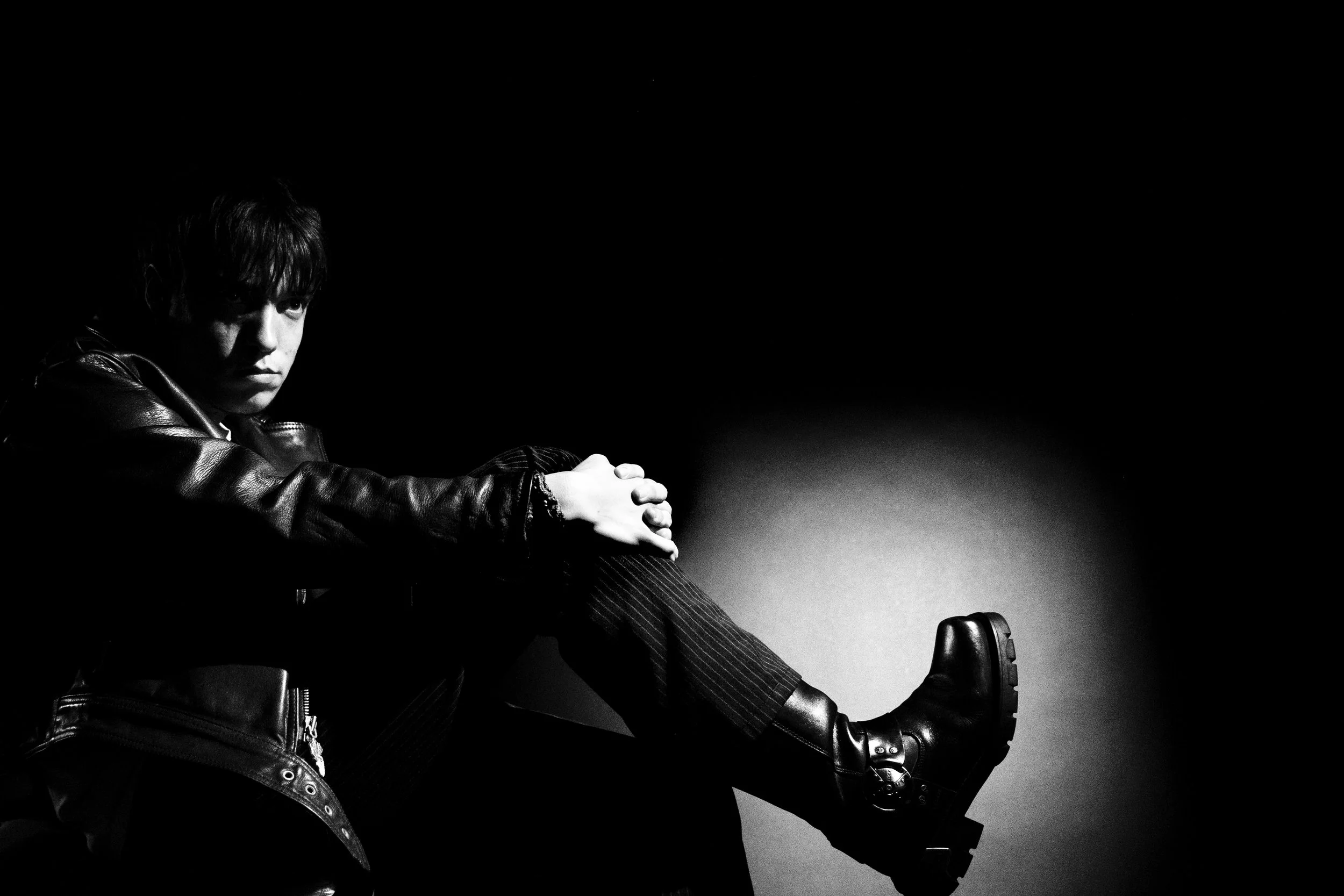Jeune homme en vêtements noirs assis dans l'obscurité. Il porte une veste en cuir, des pantalons rayés et des bottes. Il regarde pensivement la caméra.