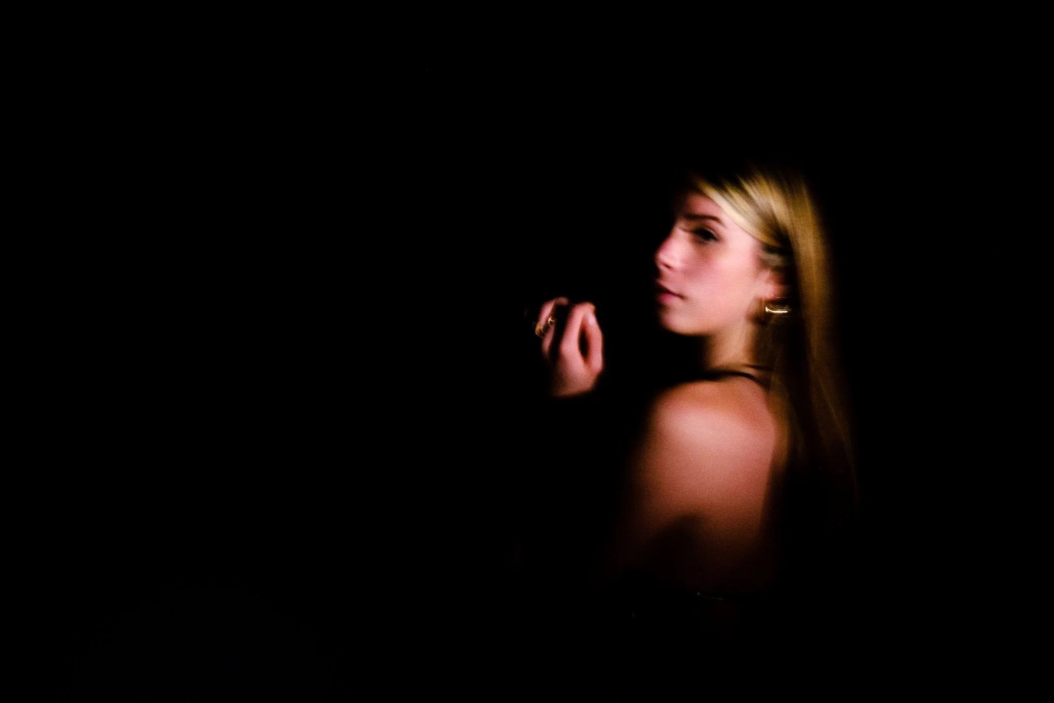 Femme aux cheveux blonds, portant des bijoux dorés, posant dans l'obscurité, éclairée partiellement.
