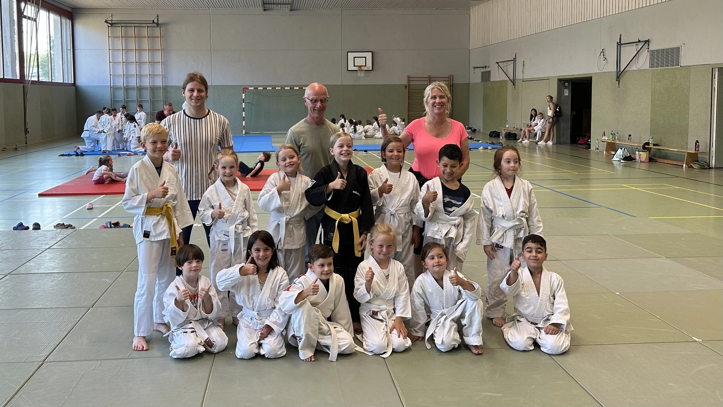 Gruppe von Kindern in Judo-Gi mit Erwachsenen in einer Turnhalle, einige Kinder tragen Gürtel in Gelb und Schwarz, sie posieren für ein Gruppenfoto, alle zeigen Daumen hoch.