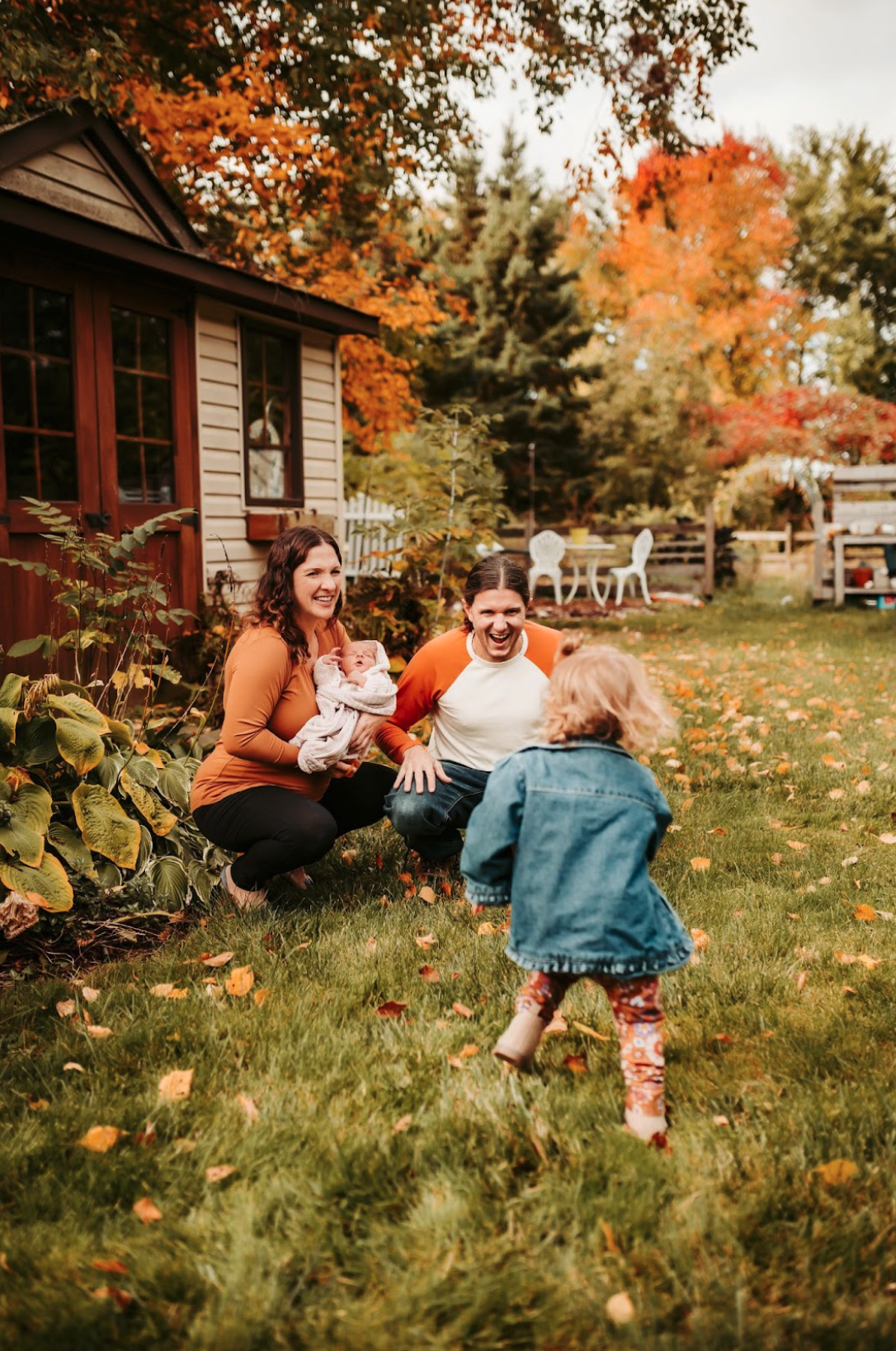Devon with her husband and children play on a grassy yard with fallen leaves, with a house, garden, and trees with autumn foliage in the background.