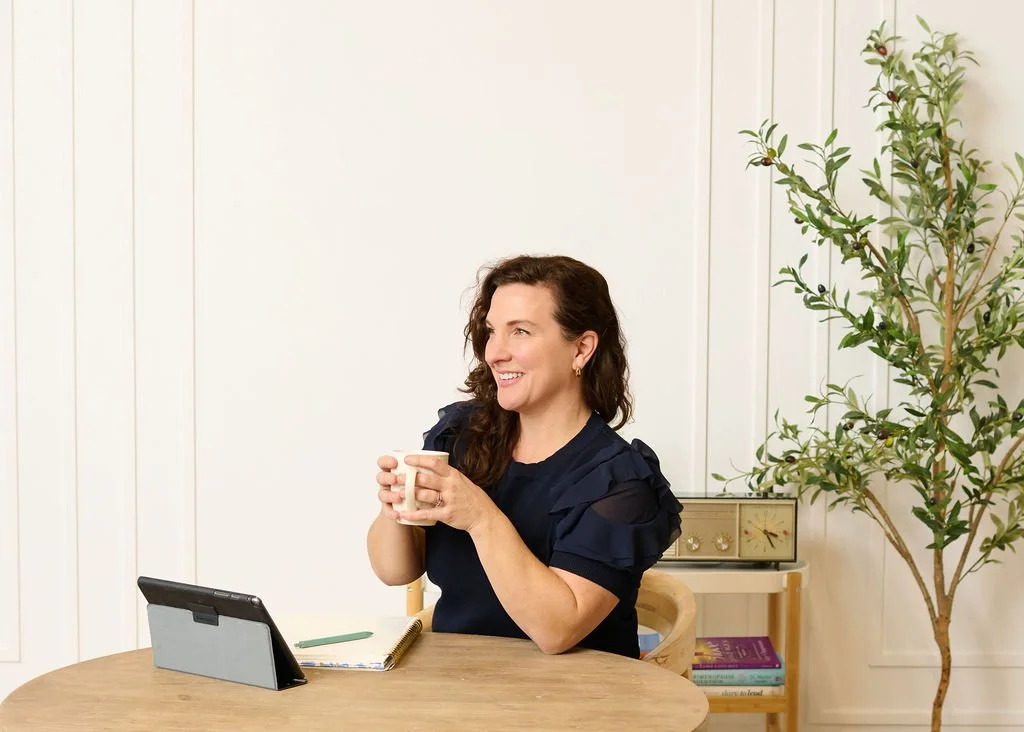 Devon with long brown hair smiling and holding a white mug while sitting at a wooden table, with a tablet, notebook, and pen in front of her, in a well-lit room with a potted plant in the background.