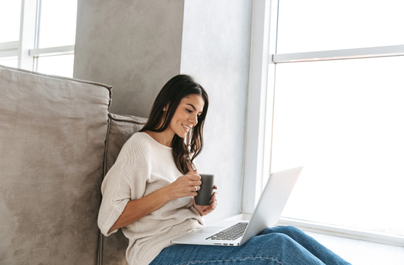 A woman sitting on a couch holding a mug and smiling while looking at her open laptop.