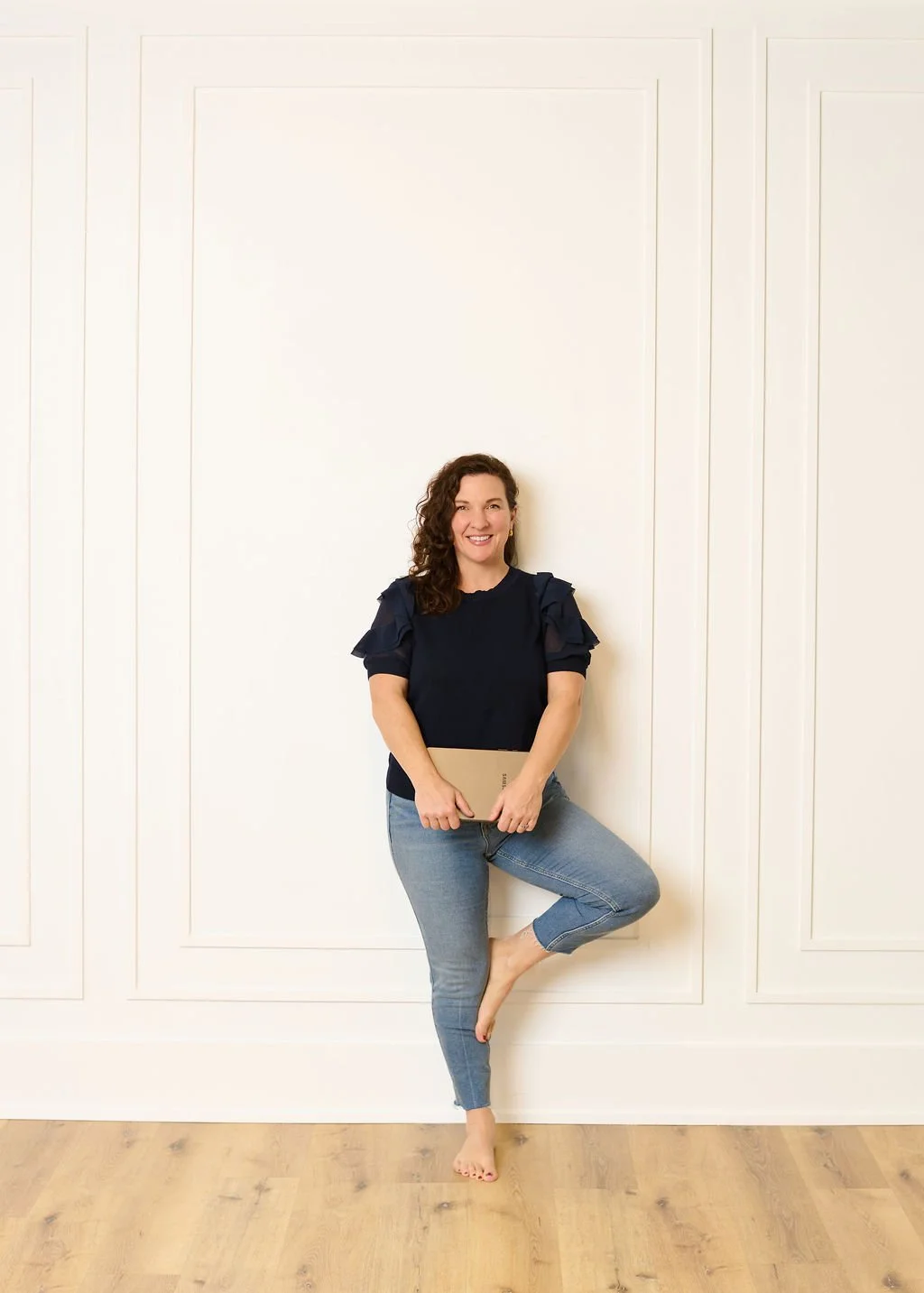 A photo of Devon with curly hair smiling, standing barefoot against a white wall with paneling, holding a tan notebook, wearing a navy blue top with ruffled sleeves and light blue jeans.