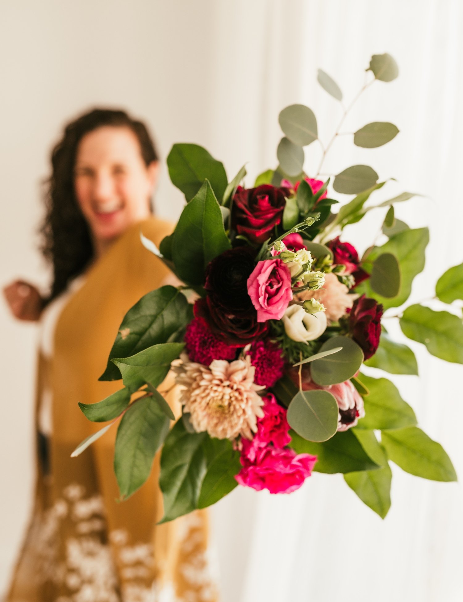 Devon with dark curly hair in a yellow jacket holding a bouquet of pink, red, and white flowers with green leaves, smiling.
