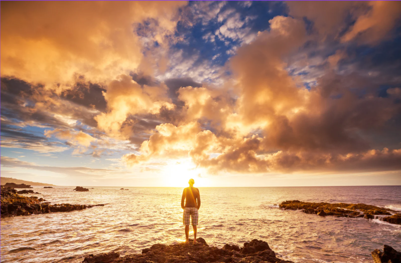 Person standing on rocks facing the ocean during a vibrant sunset with dramatic clouds