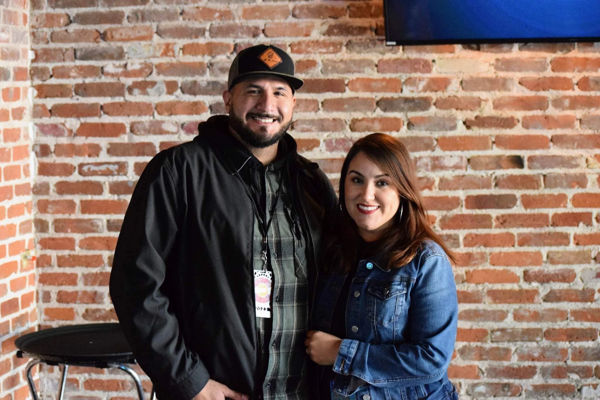 A man and woman standing close together and smiling in front of a brick wall.