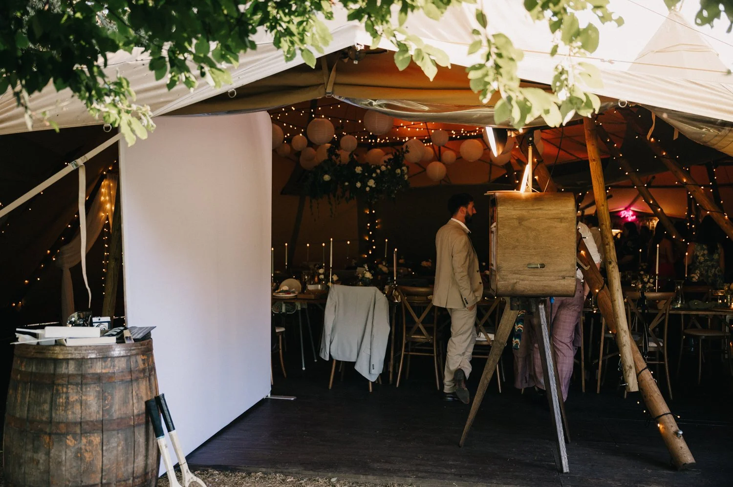 Wedding reception tent decorated with string lights, paper lanterns, and floral arrangements, featuring dining tables with candles and chairs, and a few guests present.