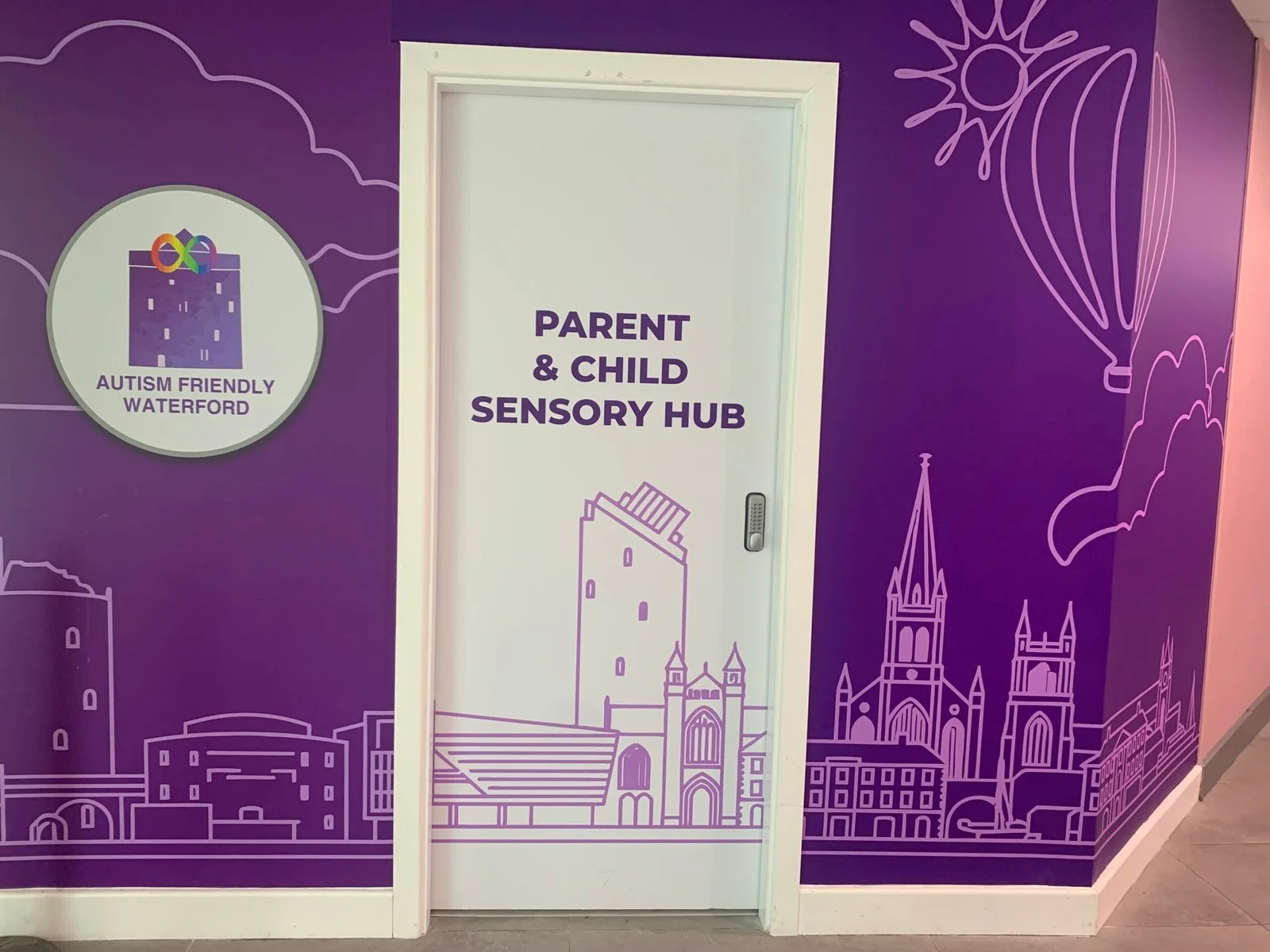 Close-up view of the white entrance door to the Parent and Child Sensory Hub with Autism Friendly Waterford branding and purple wall mural.