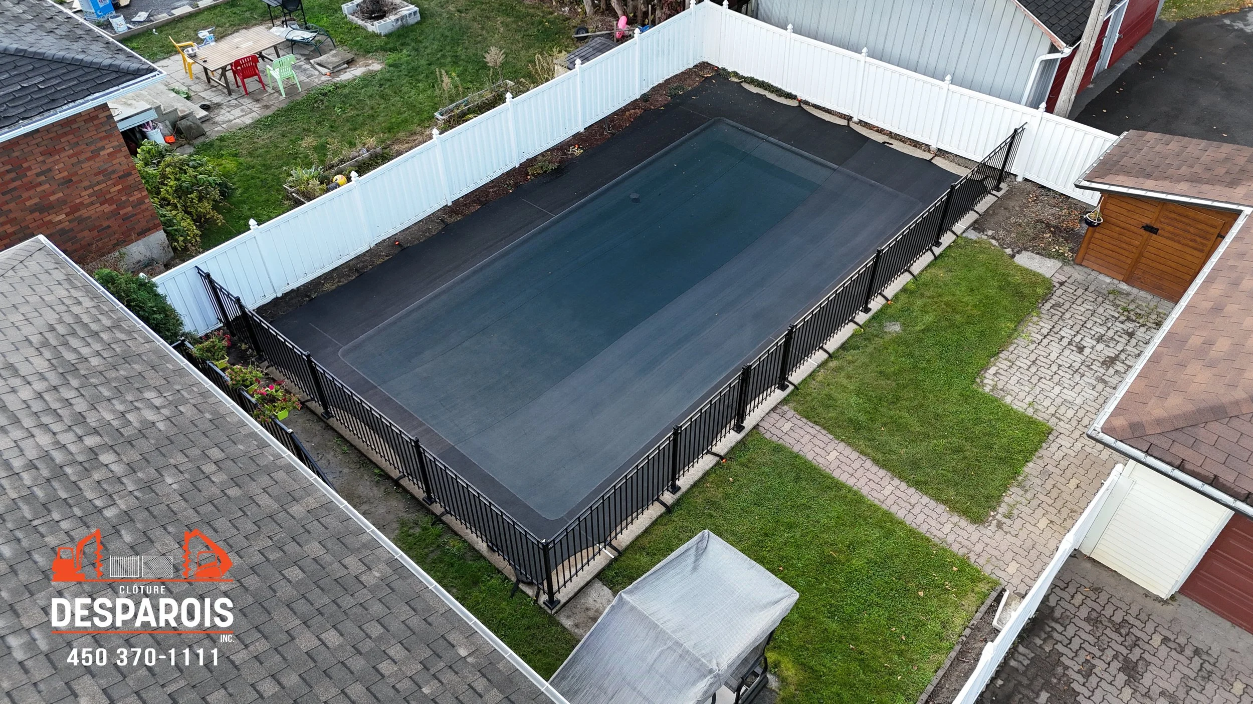 Vue aérienne d'une piscine recouverte par une couverture, entourée d'une clôture noire et blanche dans un zoo résidentiel. Proche, un petit jardin, une allée pavée, et diverses maisons adjacentes.