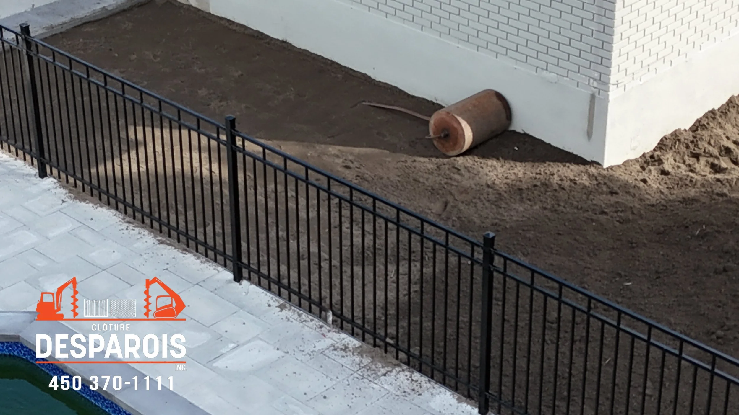 Un terrain de jardin en travaux avec une clôture en métal noir, une allée pavée et un mur de fond blanc avec des briques visibles. À côté, un tuyau enroulé rouillé repose sur le sol.