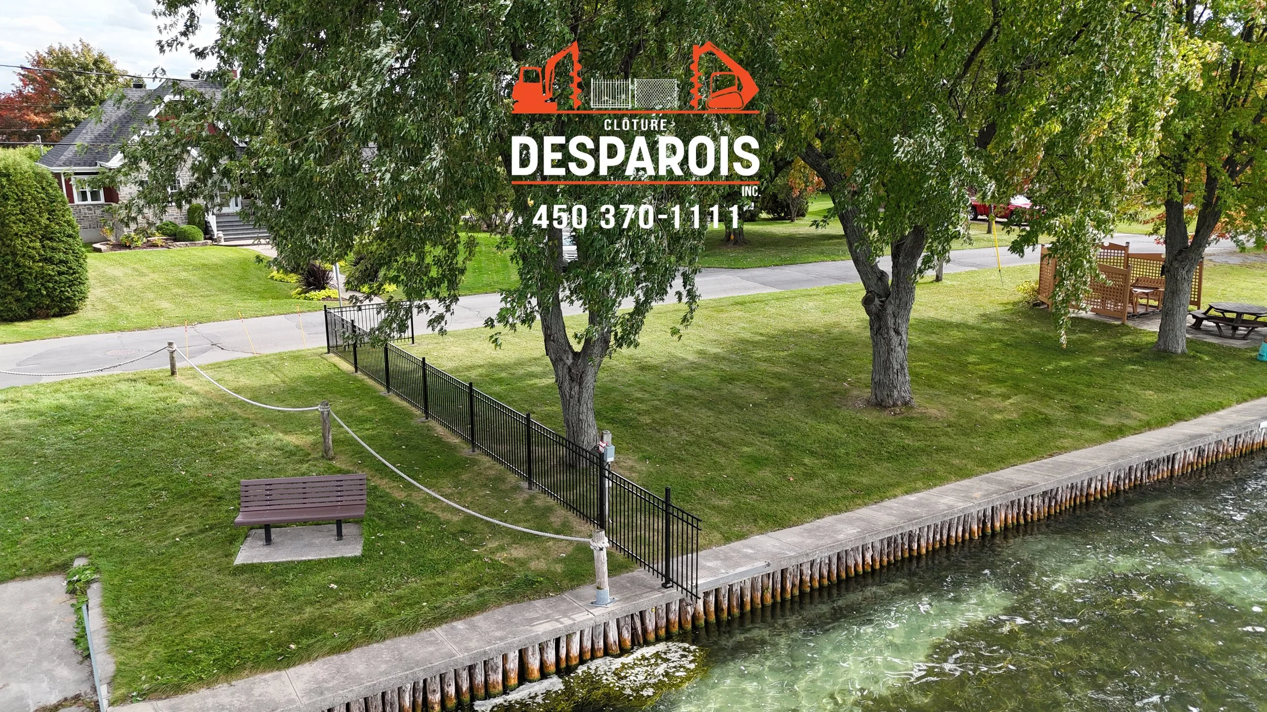 Une petite parcelle de terrain avec des arbres, une pelouse bien entretenue, un banc et une clôture, le tout à proximité d'une rivière ou d'un étang, avec une maison en arrière-plan et un logo affiché au sommet de l'image.