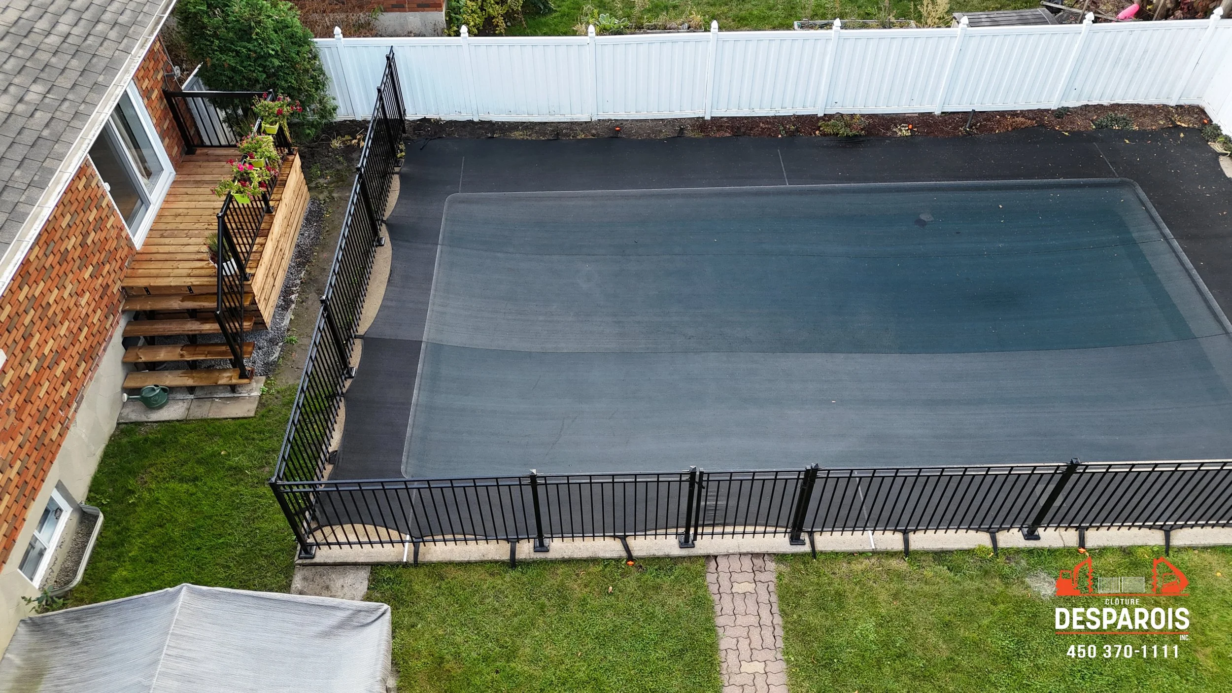 Une piscine couverte par une toile de sécurité, entourée d'une clôture noire en fer, avec un espace vert en dessous. Un patio avec un escalier en bois et des plantes en pot est visible à gauche, et un grand bâtiment en briques avec des fenêtres est a