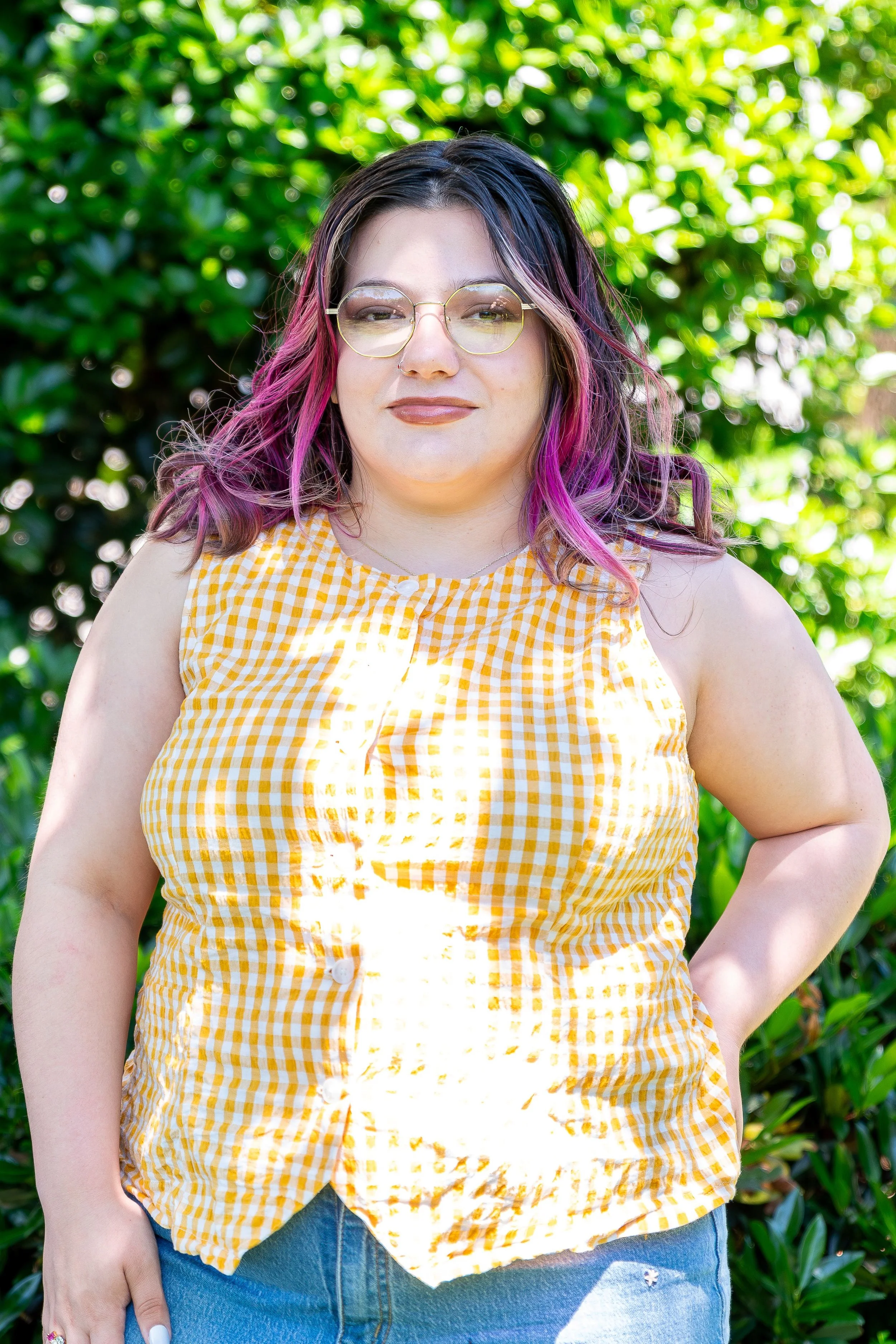 A woman with glasses and pink and purple hair standing outdoors in front of green foliage, wearing a yellow and white checkered sleeveless blouse and blue denim jeans.