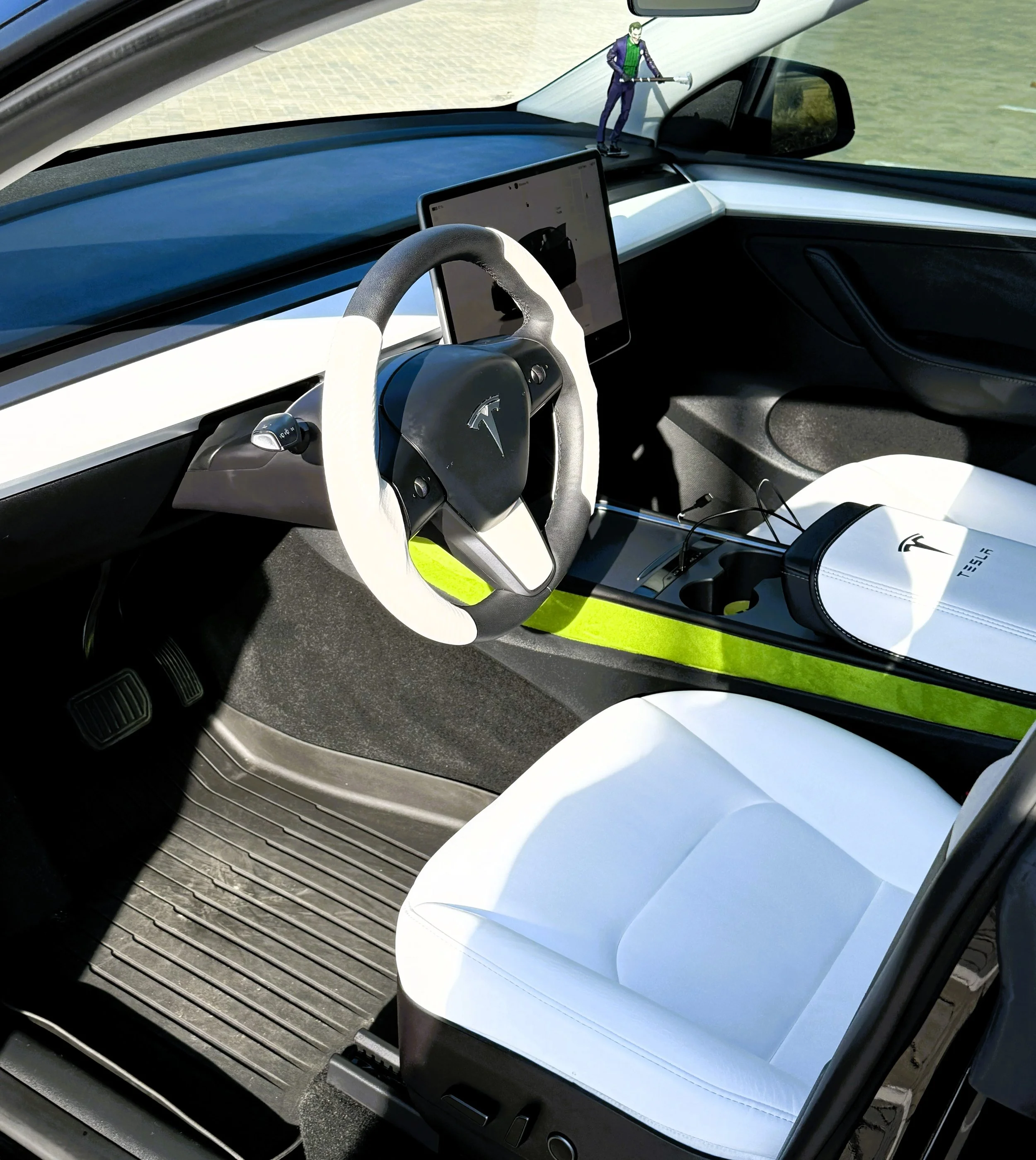 Interior of a Tesla electric vehicle with a white steering wheel cover, a white seat, and a small figure of the Joker standing on the dashboard.