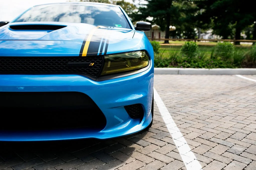 Close-up of a blue sports car parked in a parking lot with brick pavement, greenery, and trees in the background.