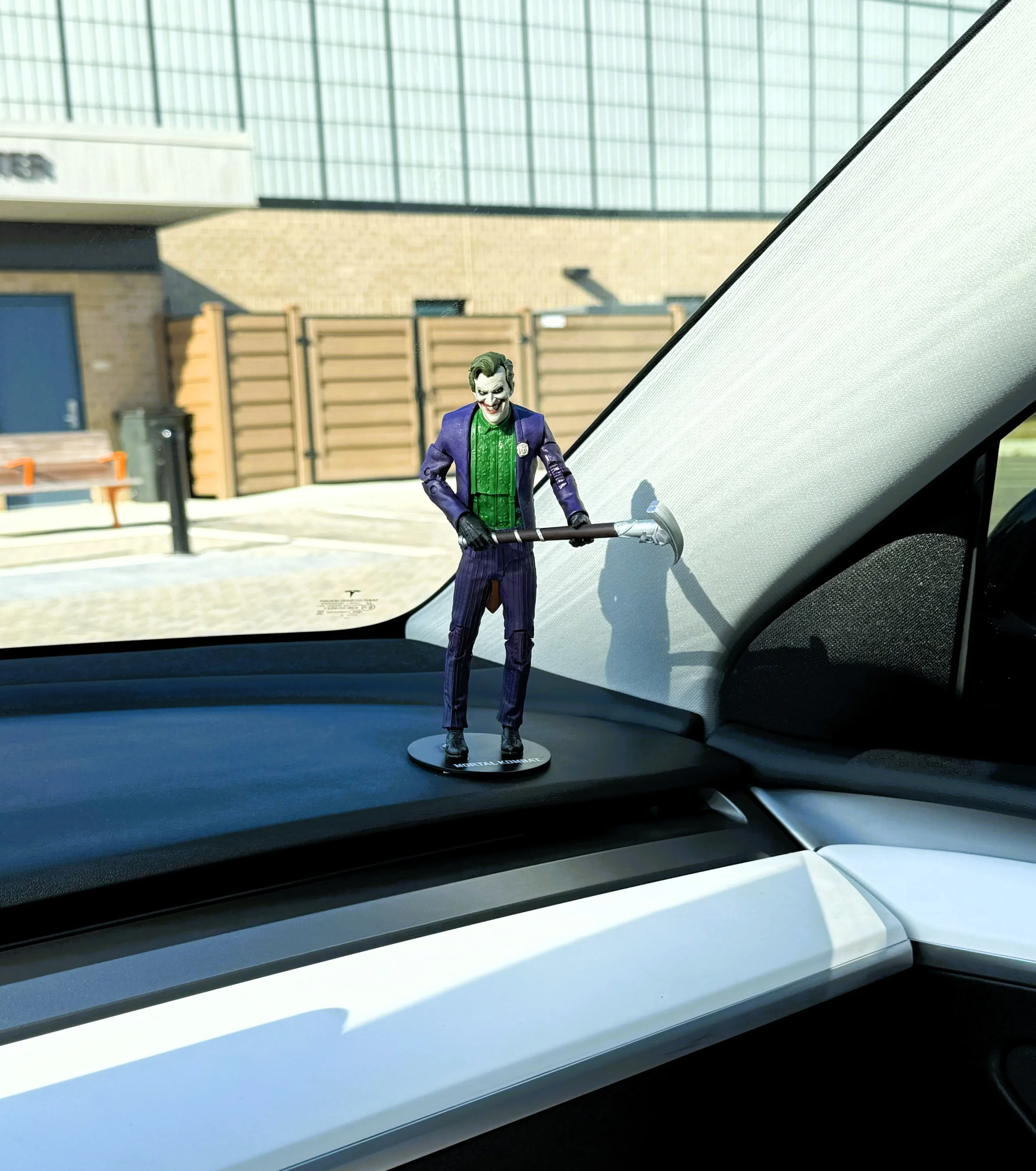 A toy figure of the Joker from Batman, dressed in a purple suit with green shirt, standing on the dashboard of a car, using a tool to clean or scrape the windshield.