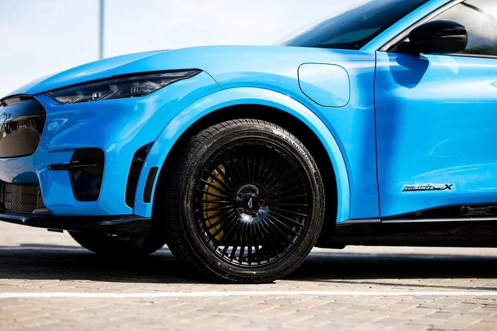 Close-up of a blue electric car's front and side, showing black wheel rims with a yellow brake caliper, side mirror, and the 'MAVERIX' badge on the side.