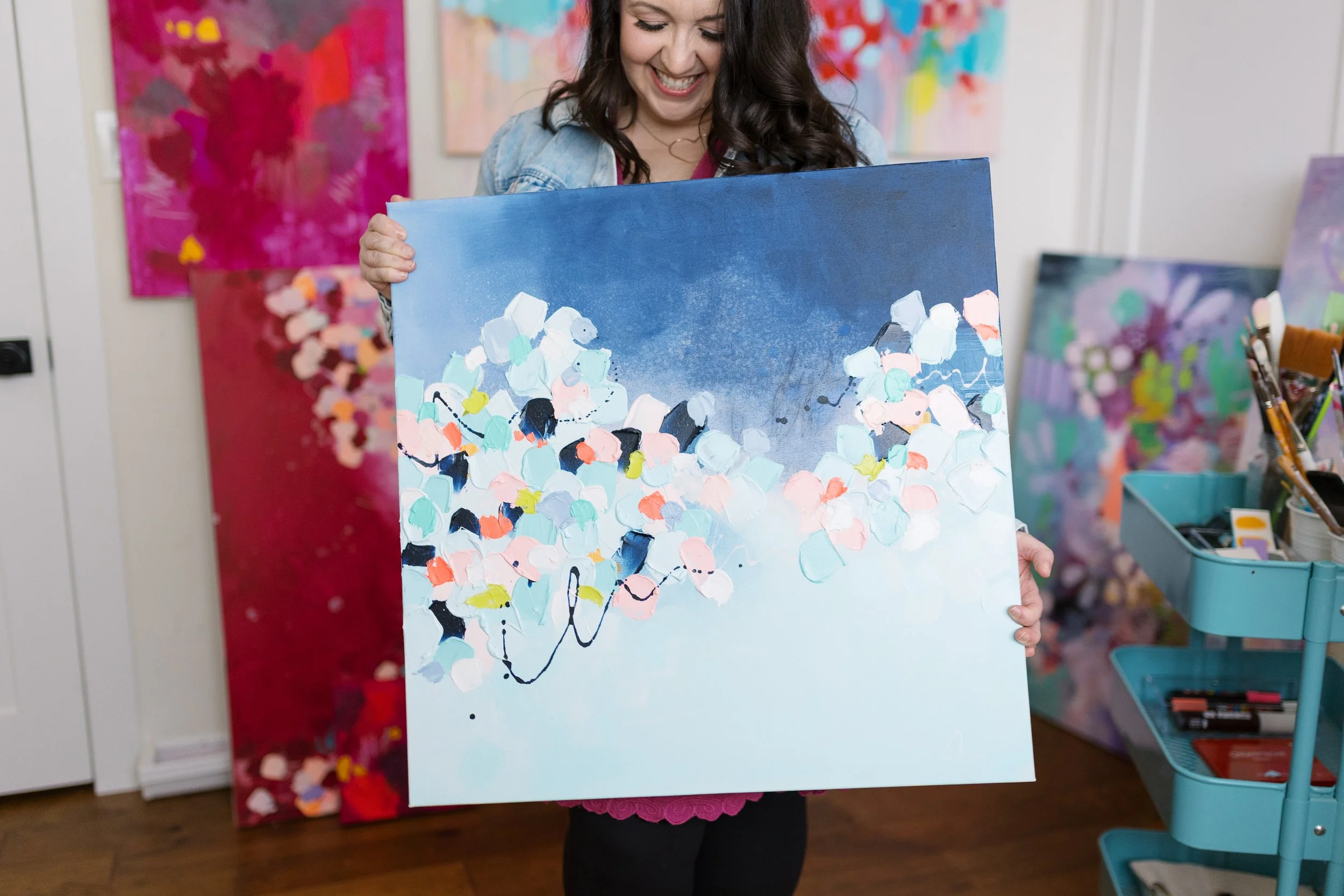 A woman holding a colorful abstract painting with a dark blue and pink background in an art studio.