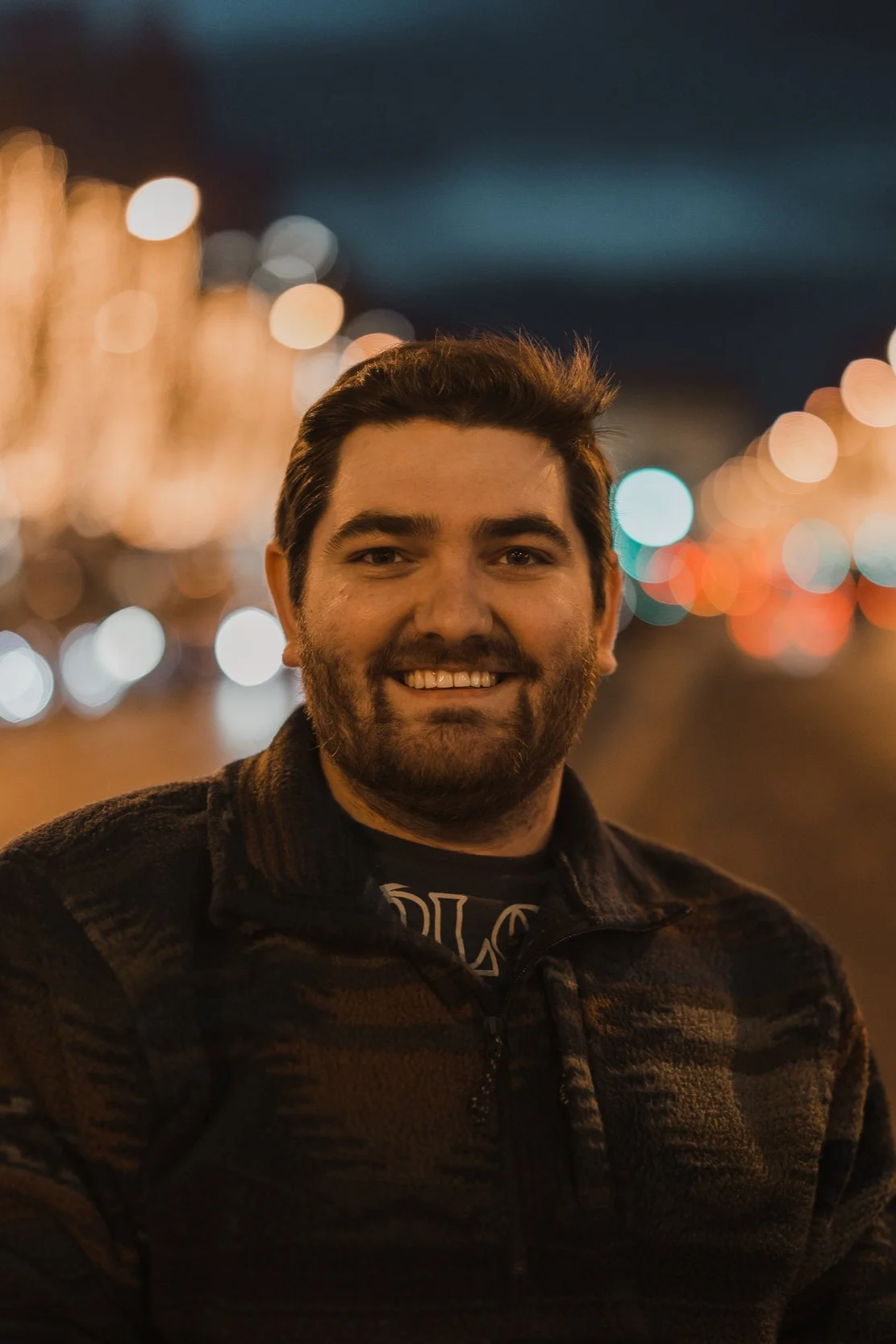 A man smiling outdoors at night with blurred city lights in the background.