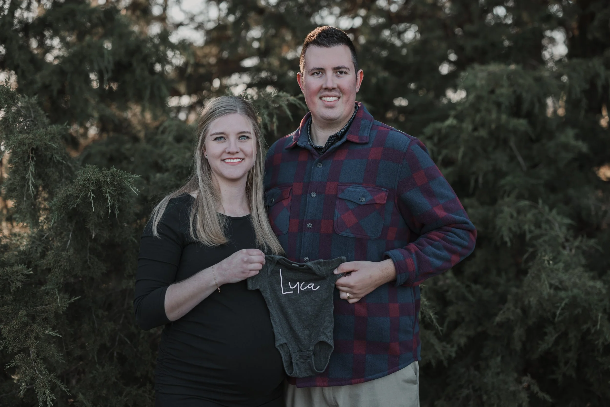 A smiling pregnant woman and a man standing outdoors in front of bushes, holding a gray baby onesie with the name 'Lyra' written on it.