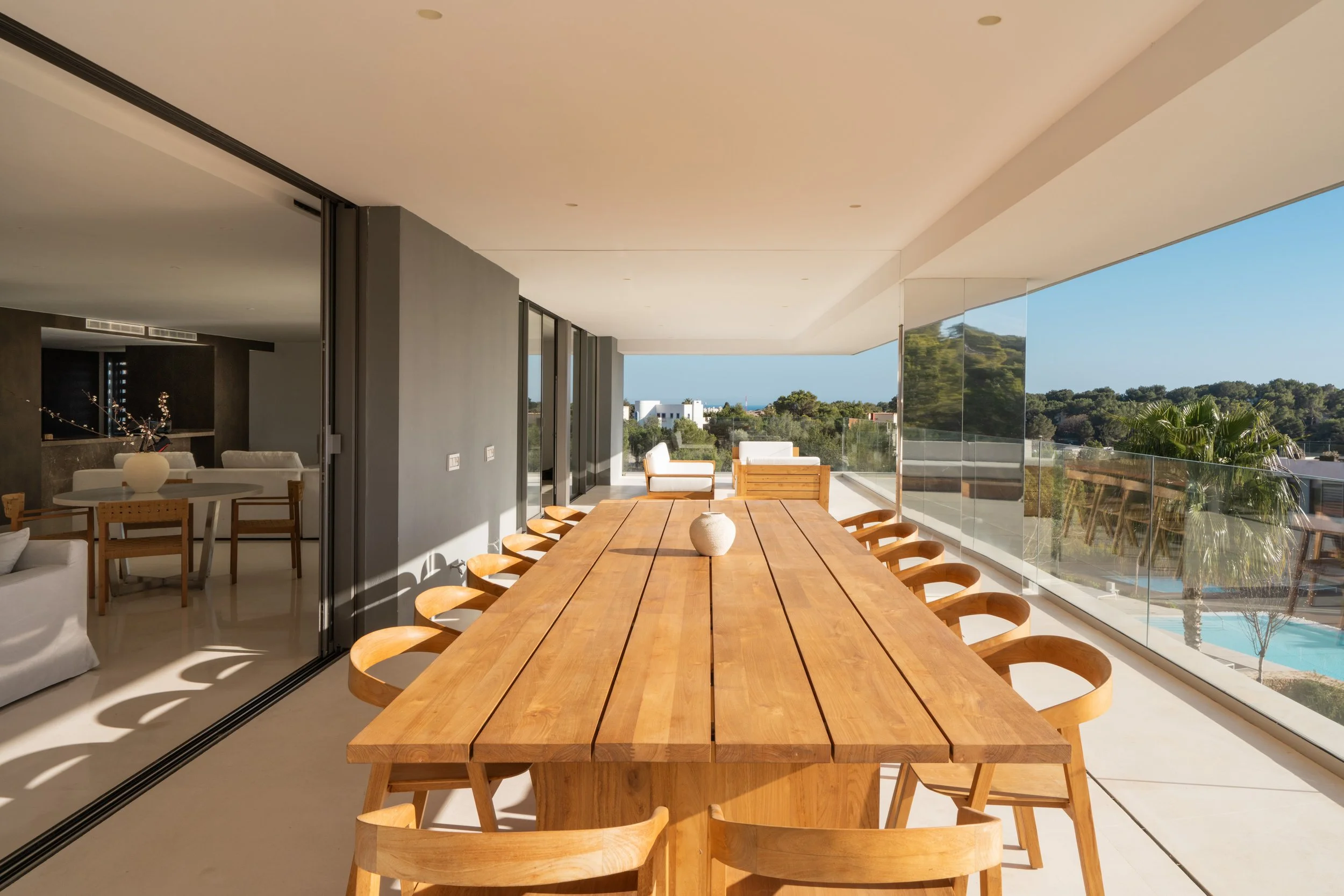 A spacious indoor-outdoor dining area with a large wooden table and chairs, overlooking a scenic landscape with trees and a swimming pool outside.