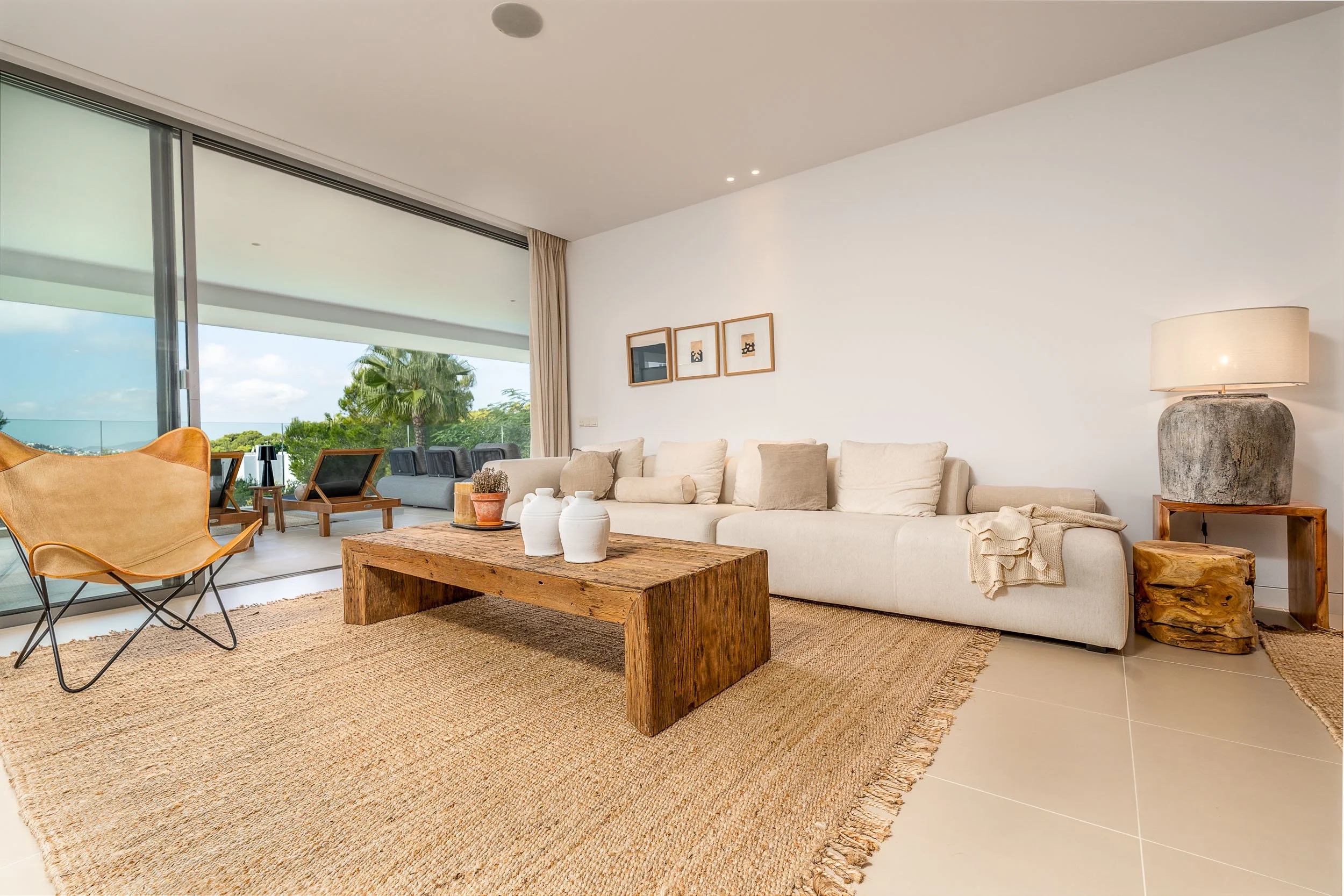 Living room with a large cream-colored sofa, a rustic wooden coffee table with white decorative vases, and a beige rug. There is a side table with a stone lamp, and artwork hanging on the white wall. Sliding glass doors open to a balcony with outdoor furniture and a view of green trees and blue sky.