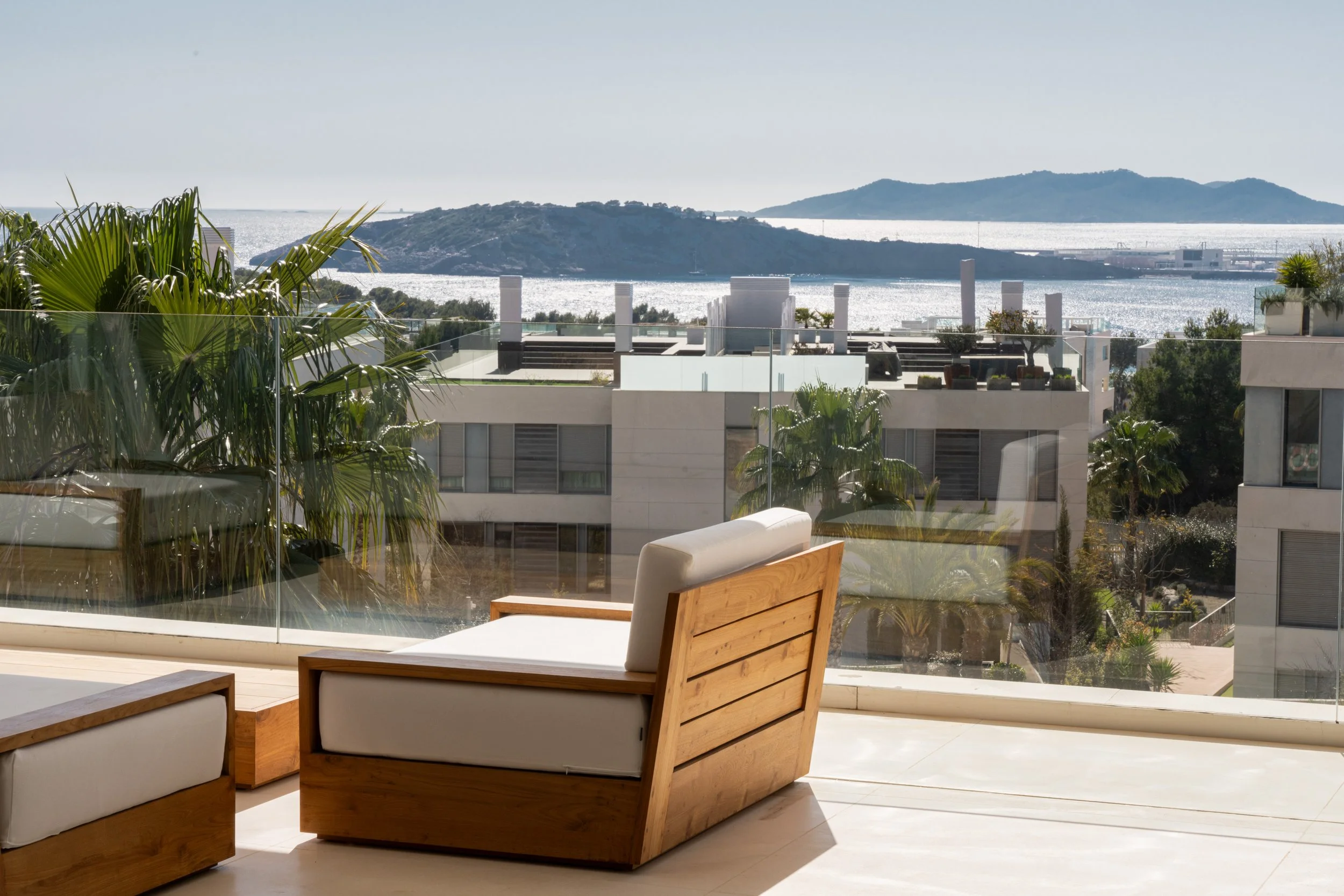 Modern balcony with wooden furniture and white cushions overlooking a cityscape, with trees, buildings, water, and hills in the background.