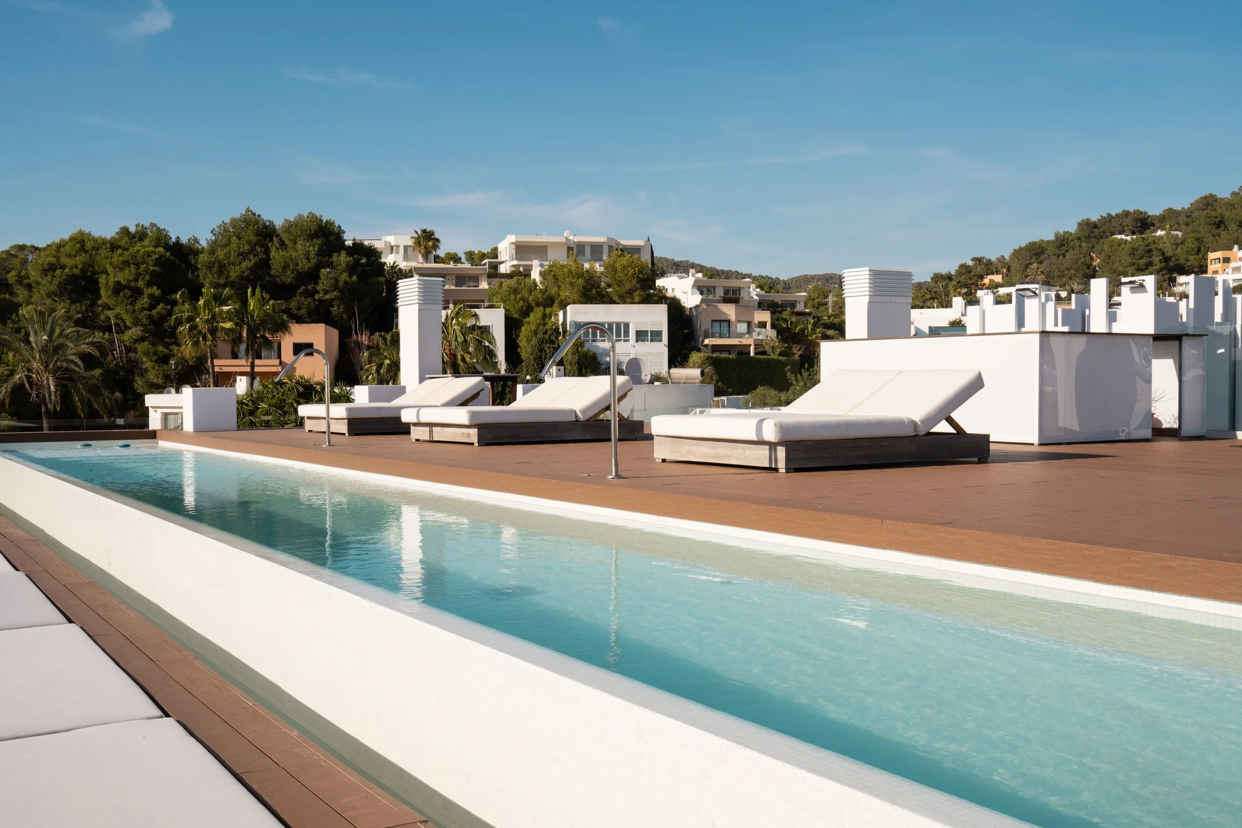 Rooftop swimming pool with white lounge chairs under a clear blue sky, overlooking a hillside community with white buildings and green trees.