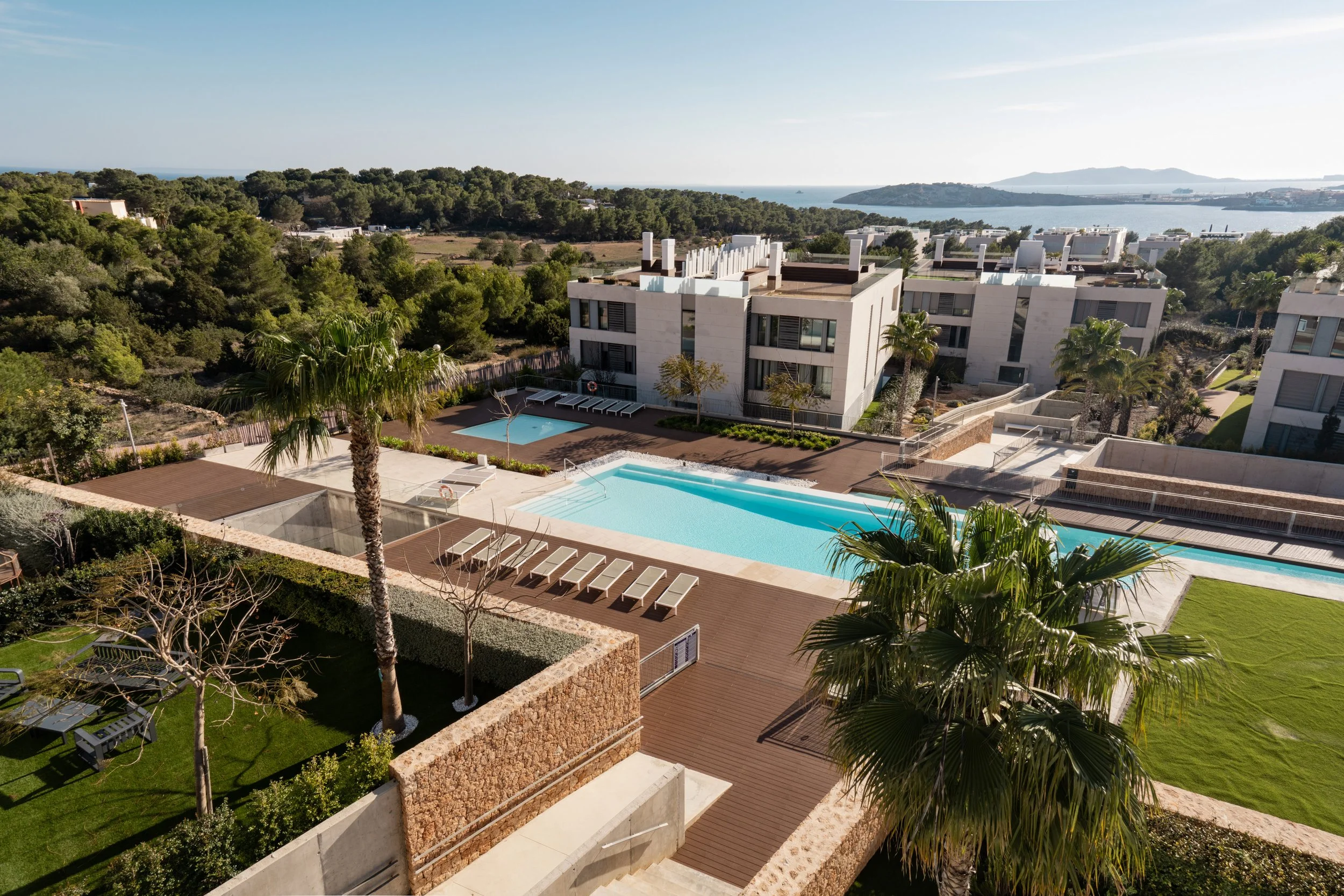 Aerial view of a modern residential complex with multiple swimming pools, palm trees, and lush green landscaping near a body of water in the background.