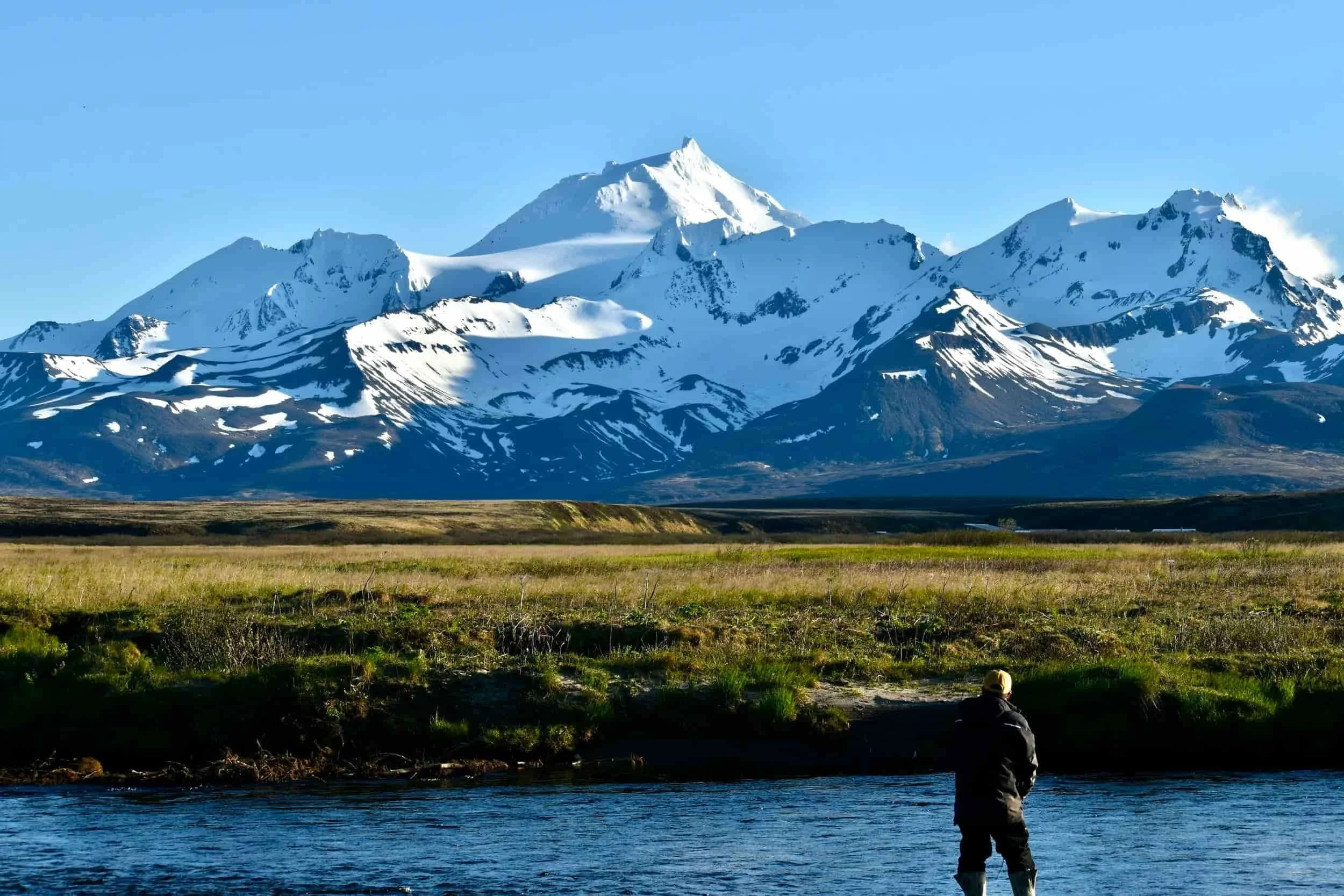 Person fishing in a river with snow-capped mountains in the background.