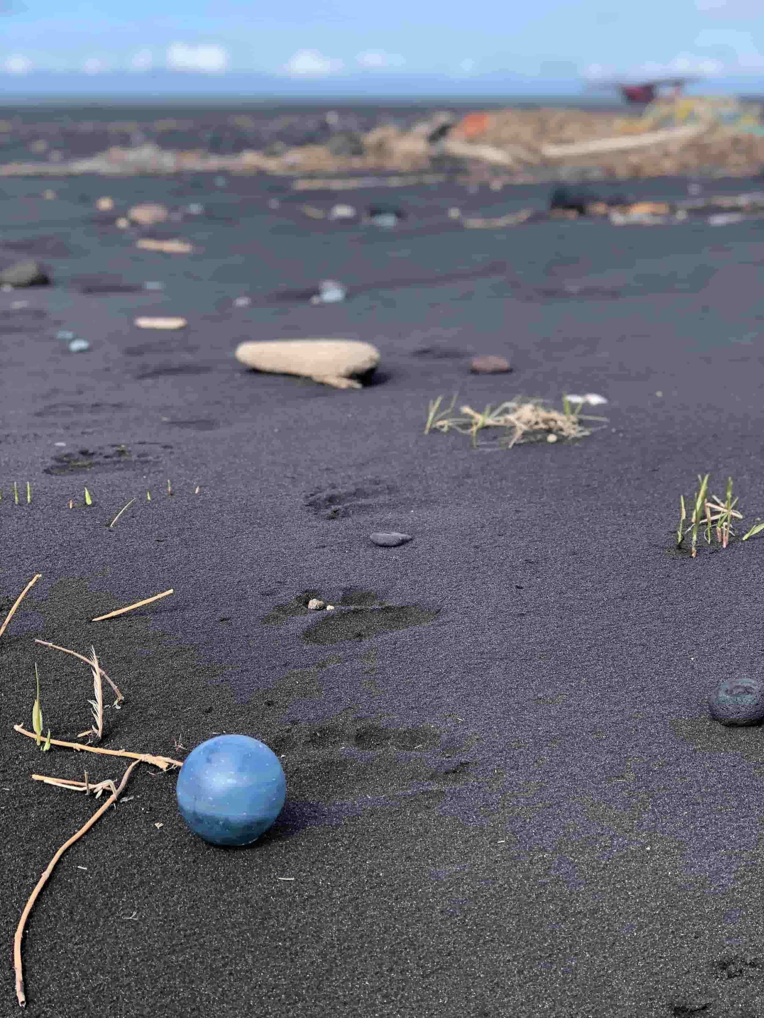 A blue marble on black volcanic sand with small rocks, footprints, and sparse grass, with a beach and airplane in the background under a partly cloudy sky.