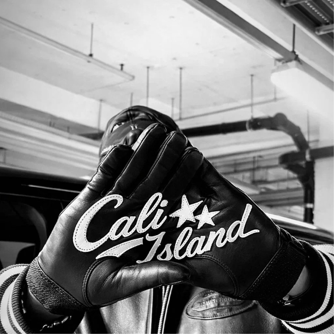 A person holding up their hand wearing a black leather glove with white script reading 'Cali Island' and two stars, in an indoor parking garage.