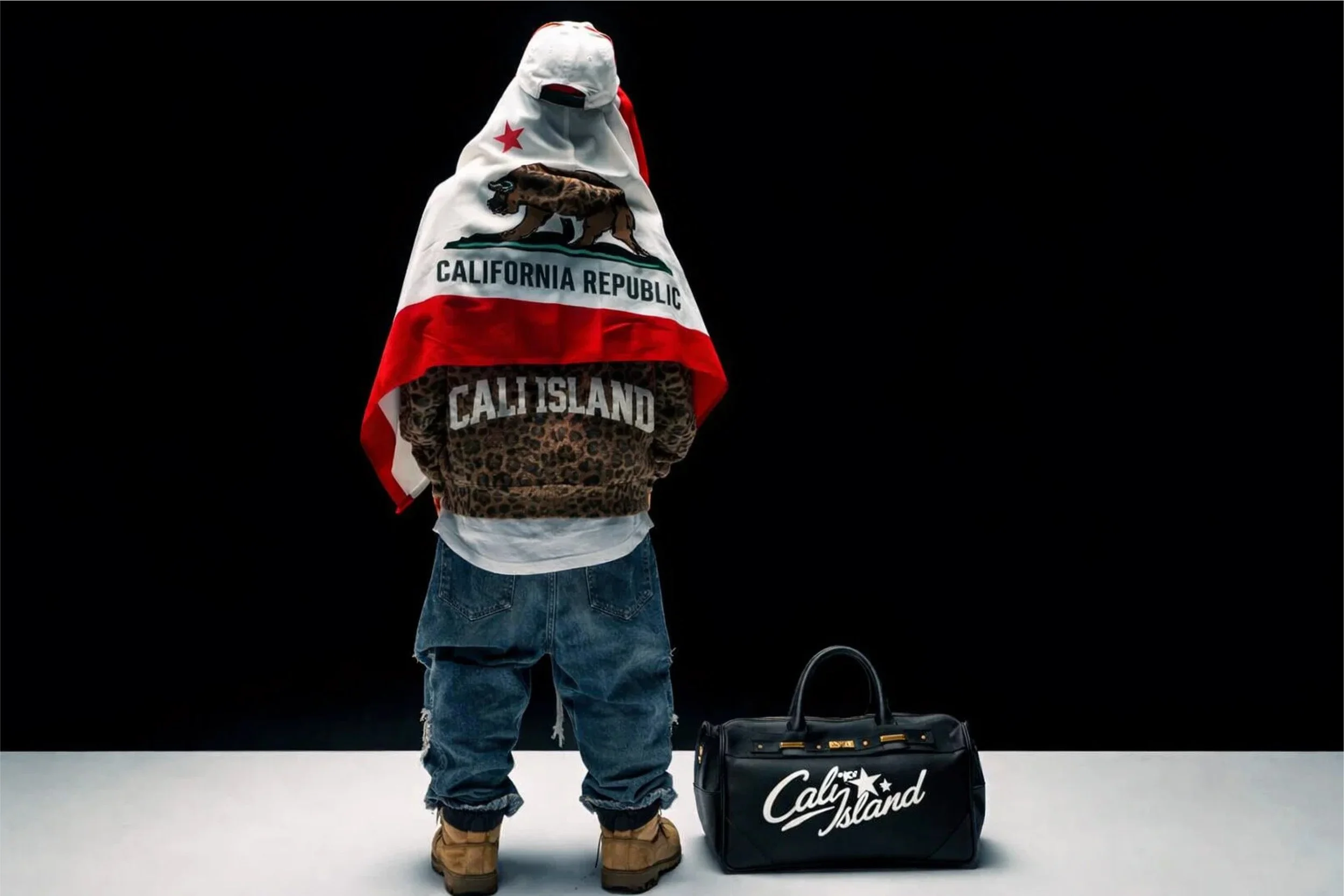 Person wearing a California Republic flag with a bear and star, a leopard print hoodie with 'CALI ISLAND' text, blue jeans, and tan boots, standing next to black Caly Island branded duffle bag against a black background.