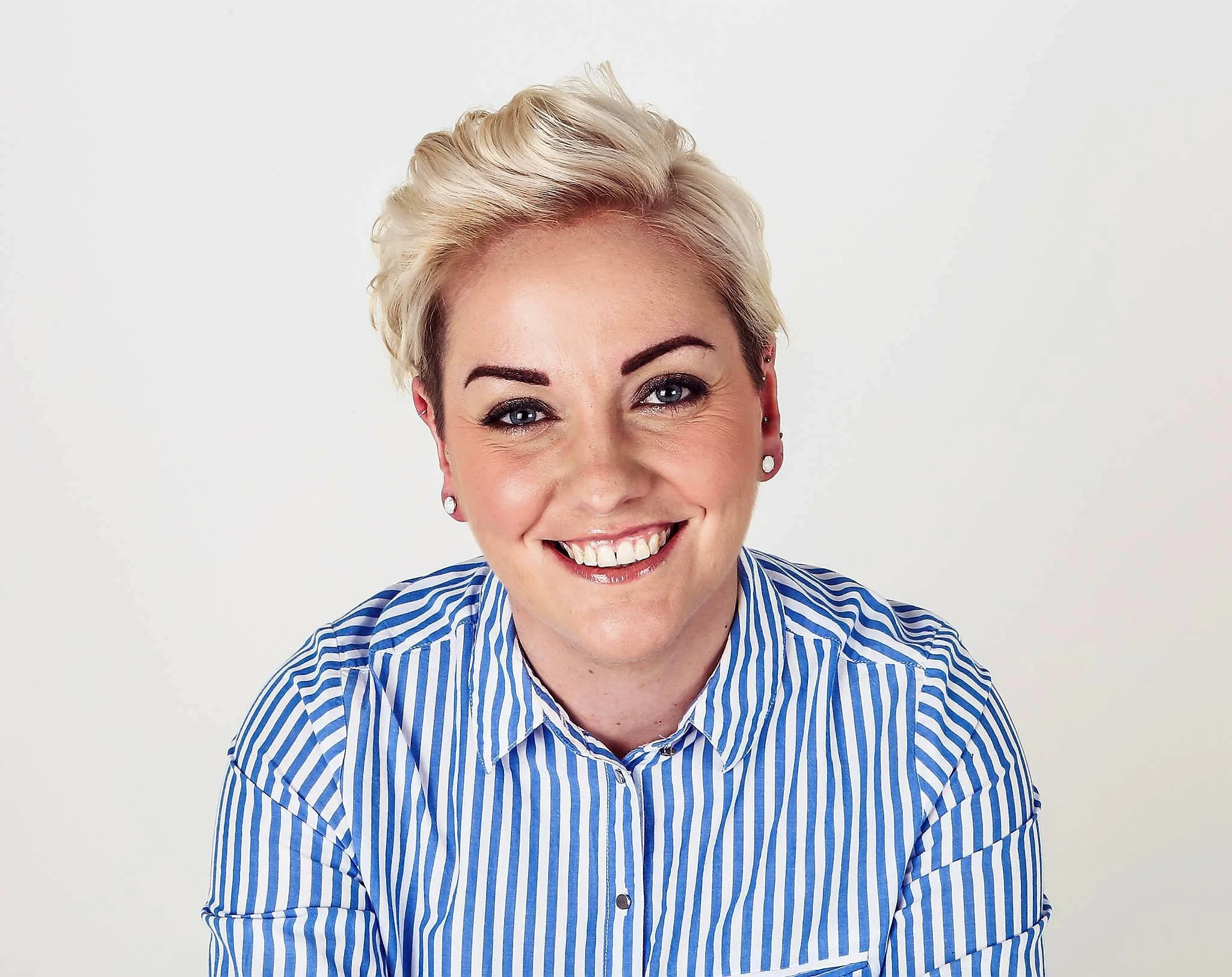 Smiling person with short blonde hair in a blue and white striped shirt against a plain background. Bright and cheerful mood.