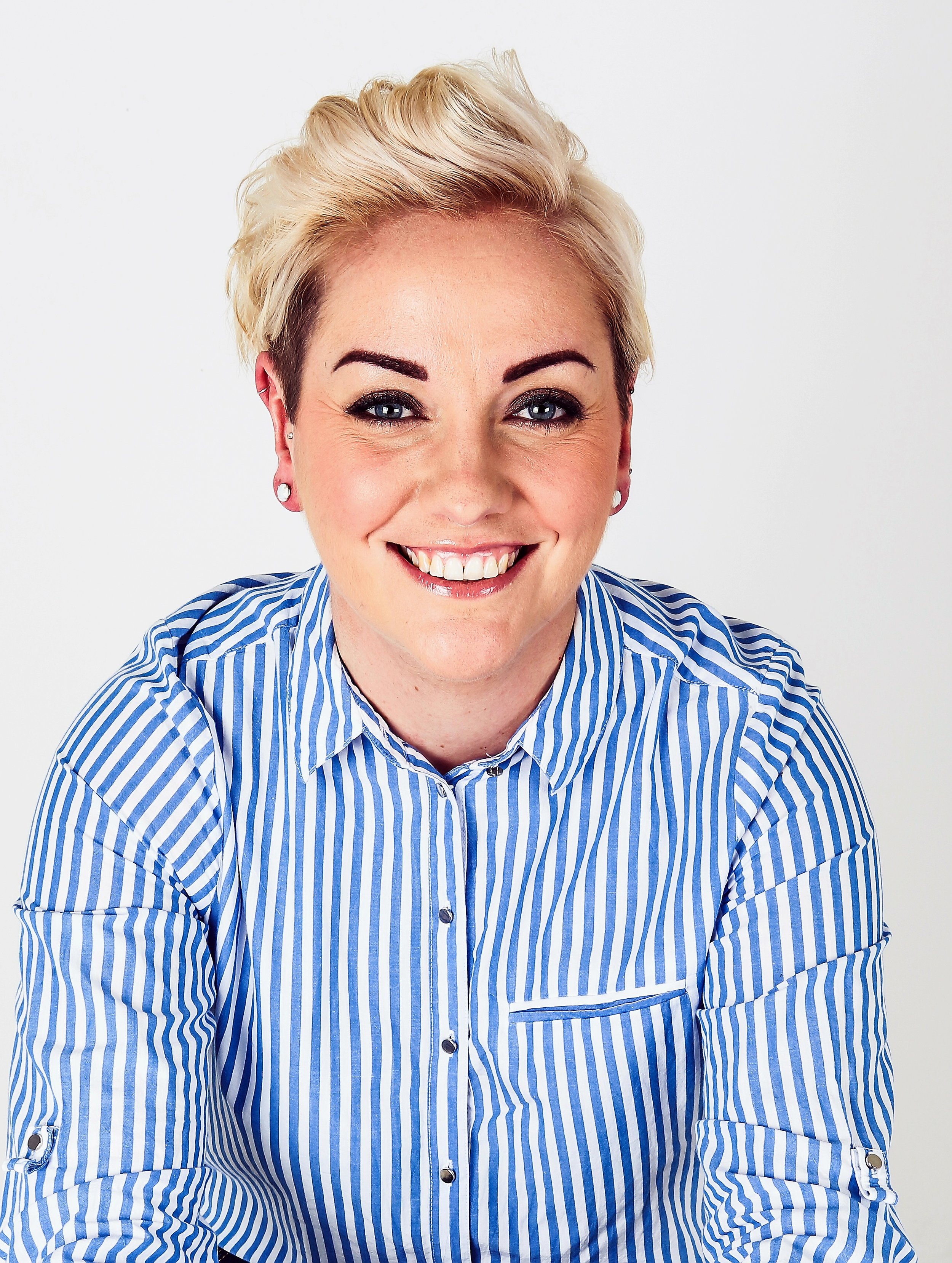 Smiling person with short blonde hair in a blue and white striped shirt against a plain background. Bright and cheerful mood.