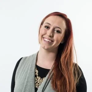 Young woman with red hair smiling, wearing a black top, a necklace, and a grey vest against a white background.