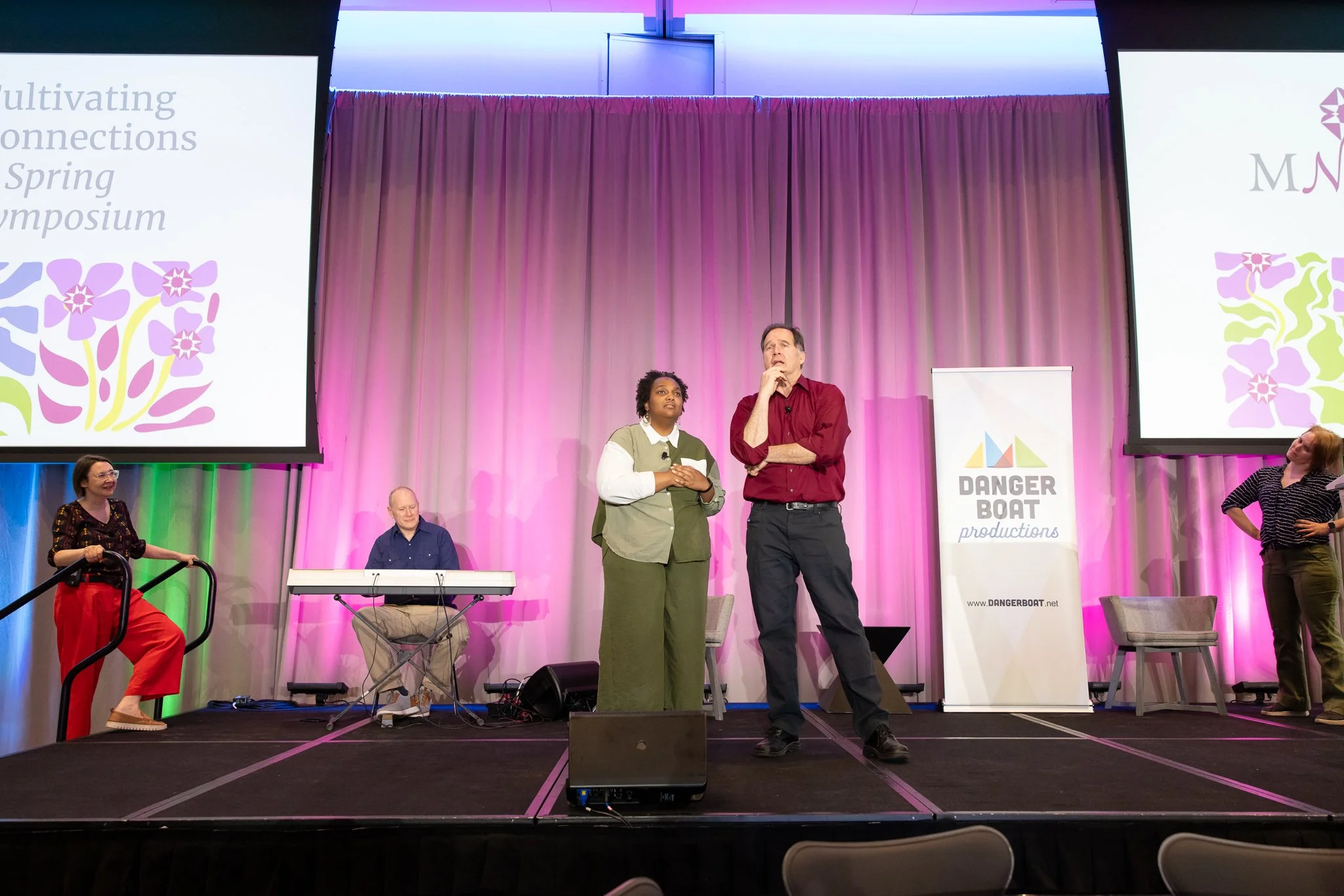 An African American person in green pants and a block patterned long-sleeve shirt stands next to a white man in black pants and a red button-up shirt onstage at a conference.