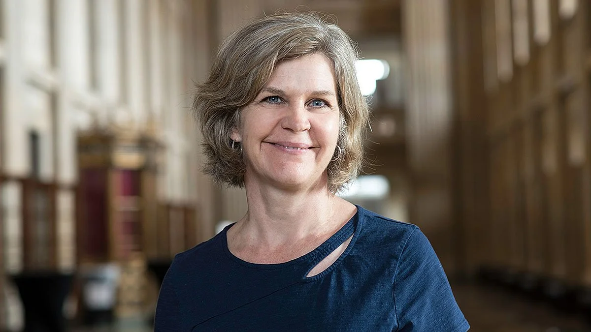 A smiling woman with shoulder-length light brown hair and blue eyes standing inside a library.