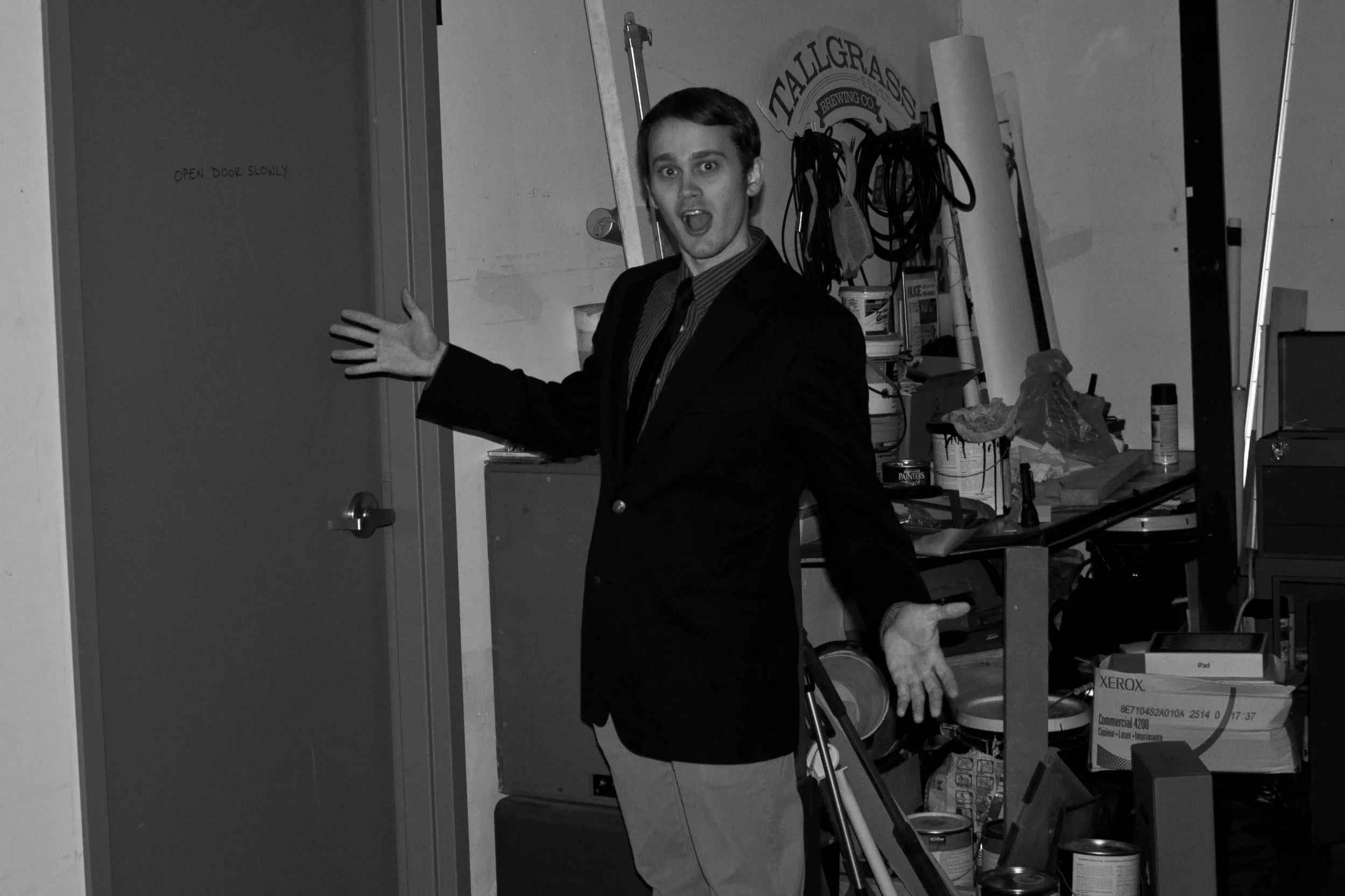 Black and white photo of a young white man with short hair in black suit jacket with tie holding his hands out to pose for a photo.