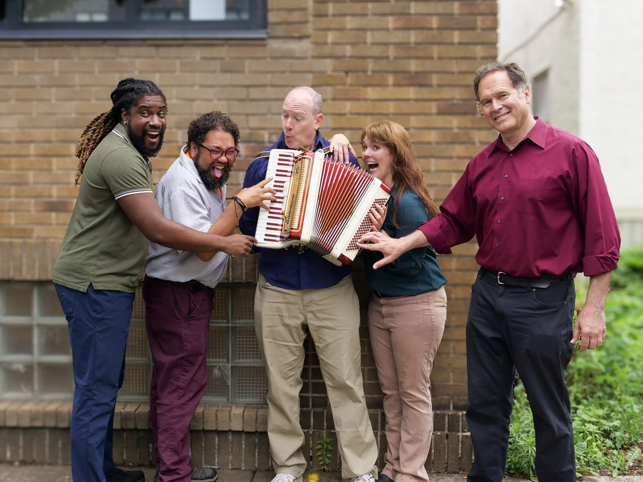 Five people standing in front of a brick wall. One man in the middle is playing the accordion while the others touch different buttons on the instrument.