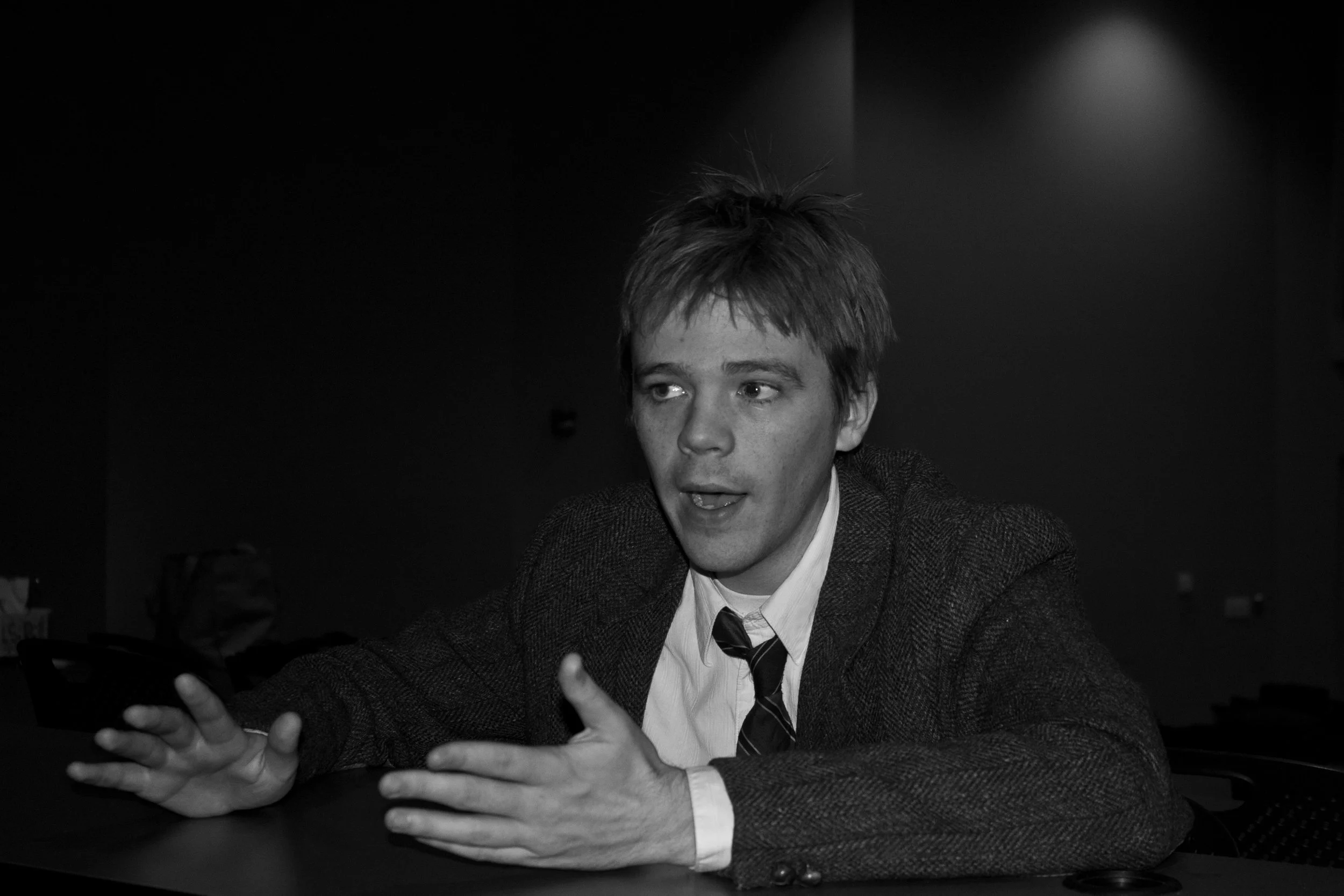 Black and white photo of a young white man in a suit jacket with a white dress shirt and a tie talking to someone out of frame.