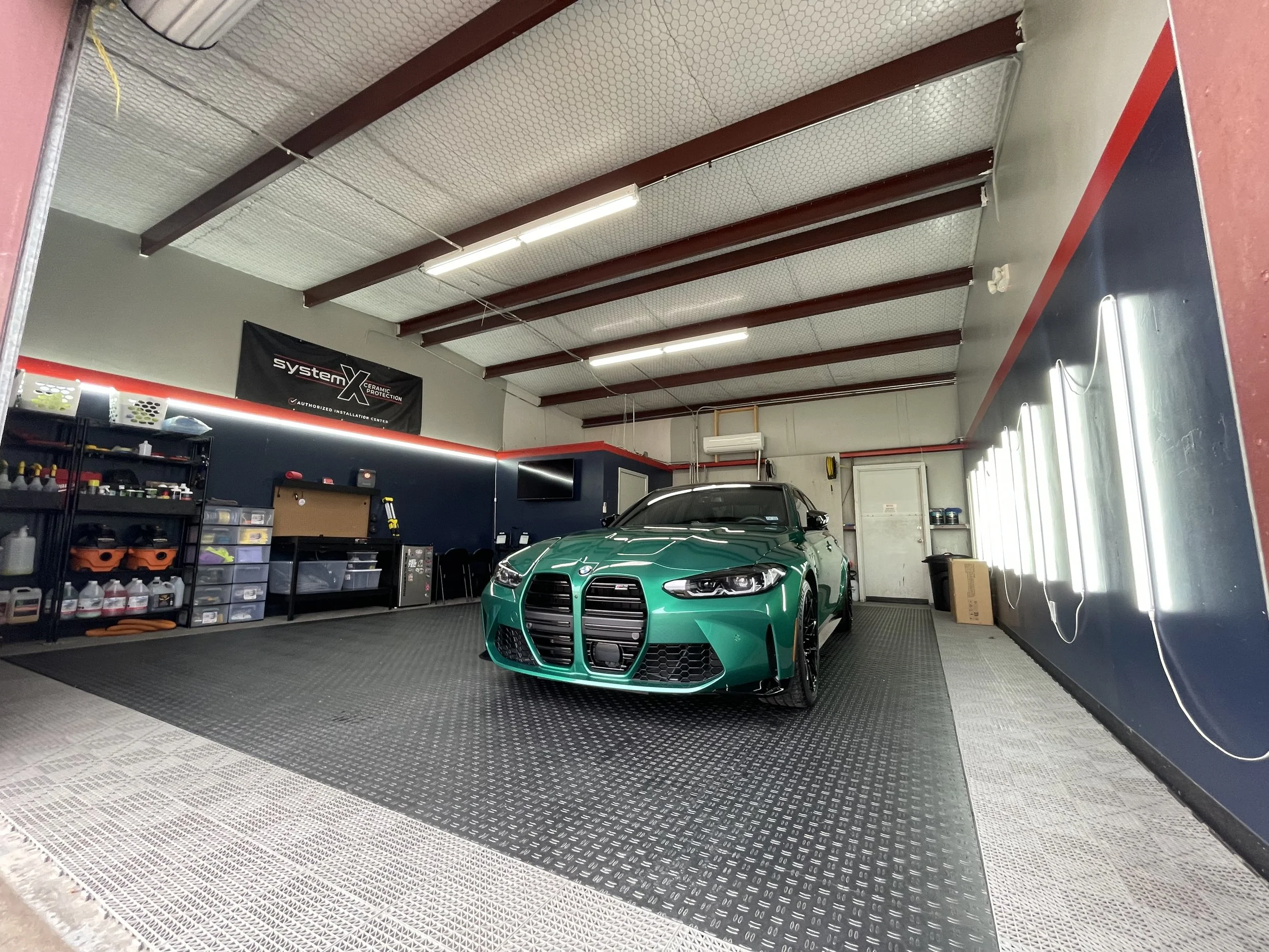 Inside a clean, well-lit garage with a green sports car parked on a black rubber flooring. The garage walls have shelves with tools and supplies, a TV mounted on the wall, and a banner that reads 'System X Ceramic Protection'. Sunlight is coming in through the windows on the right side.