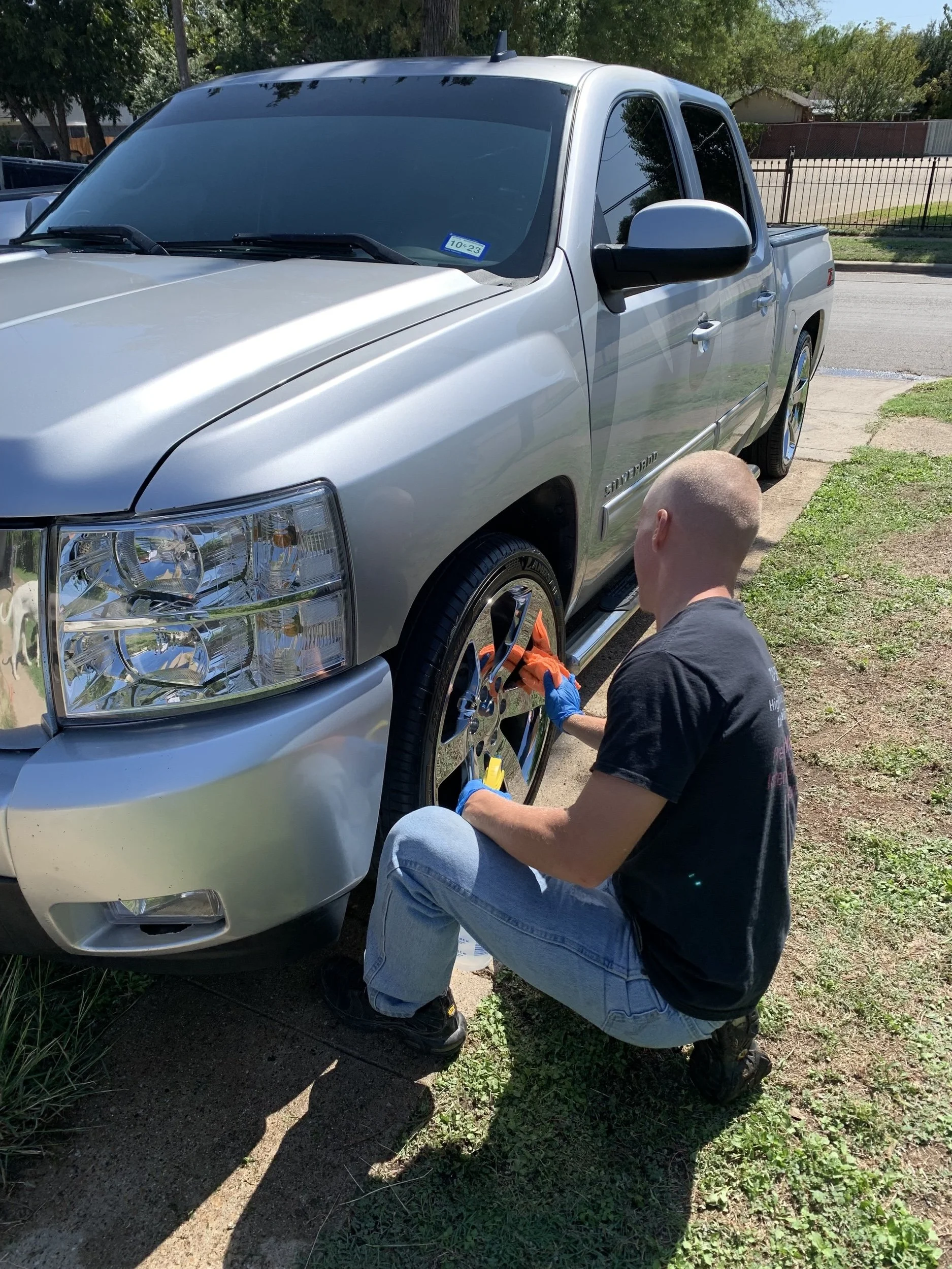 A man kneeling beside a silver pickup truck, cleaning or polishing the front left wheel on a sunny day.