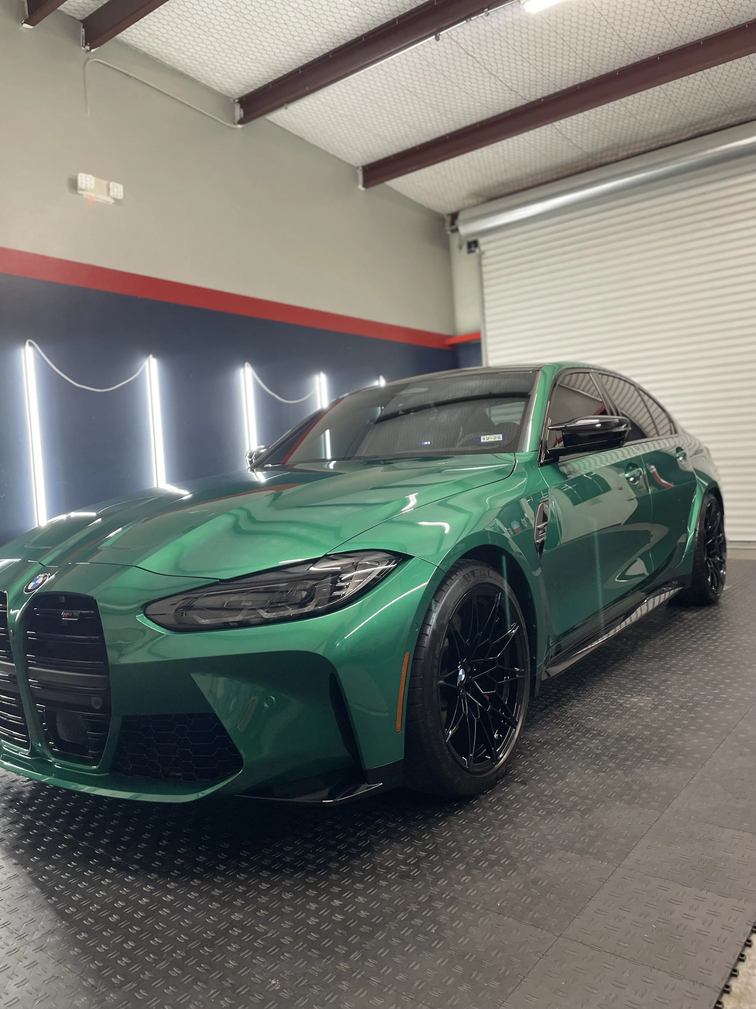 A green BMW car parked indoors on a dark textured floor with a blue and gray wall and white neon lights in the background.