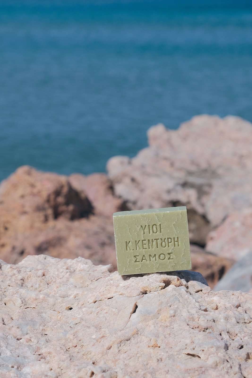 Grön tvål med grekisk text placerad på stenar vid havet.
