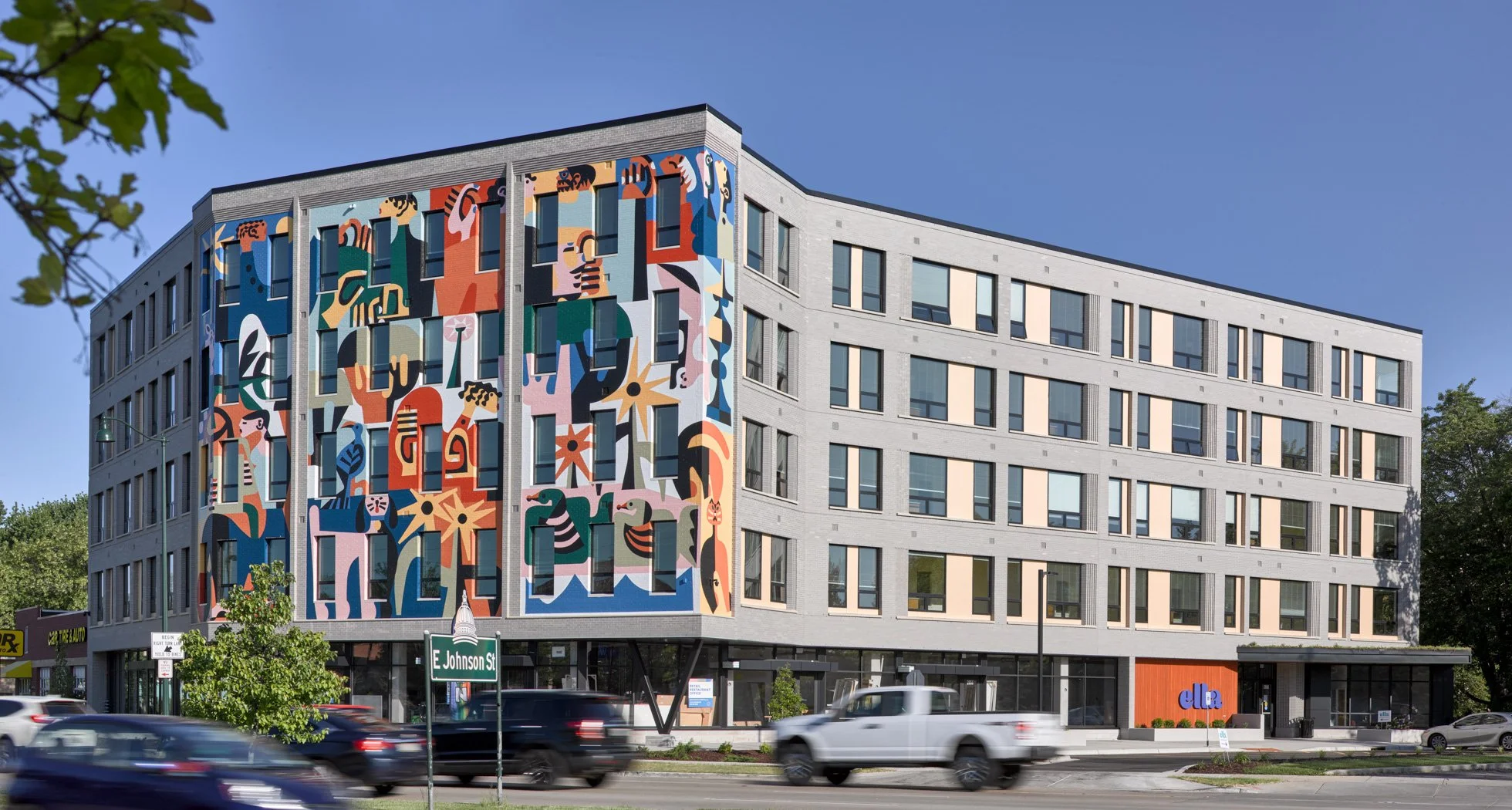 Multi-color mural facade of an apartment building, Ella Apartments. Figures in the mural include flowers, animals, and a diverse group of people. Cars drive by in the foreground.